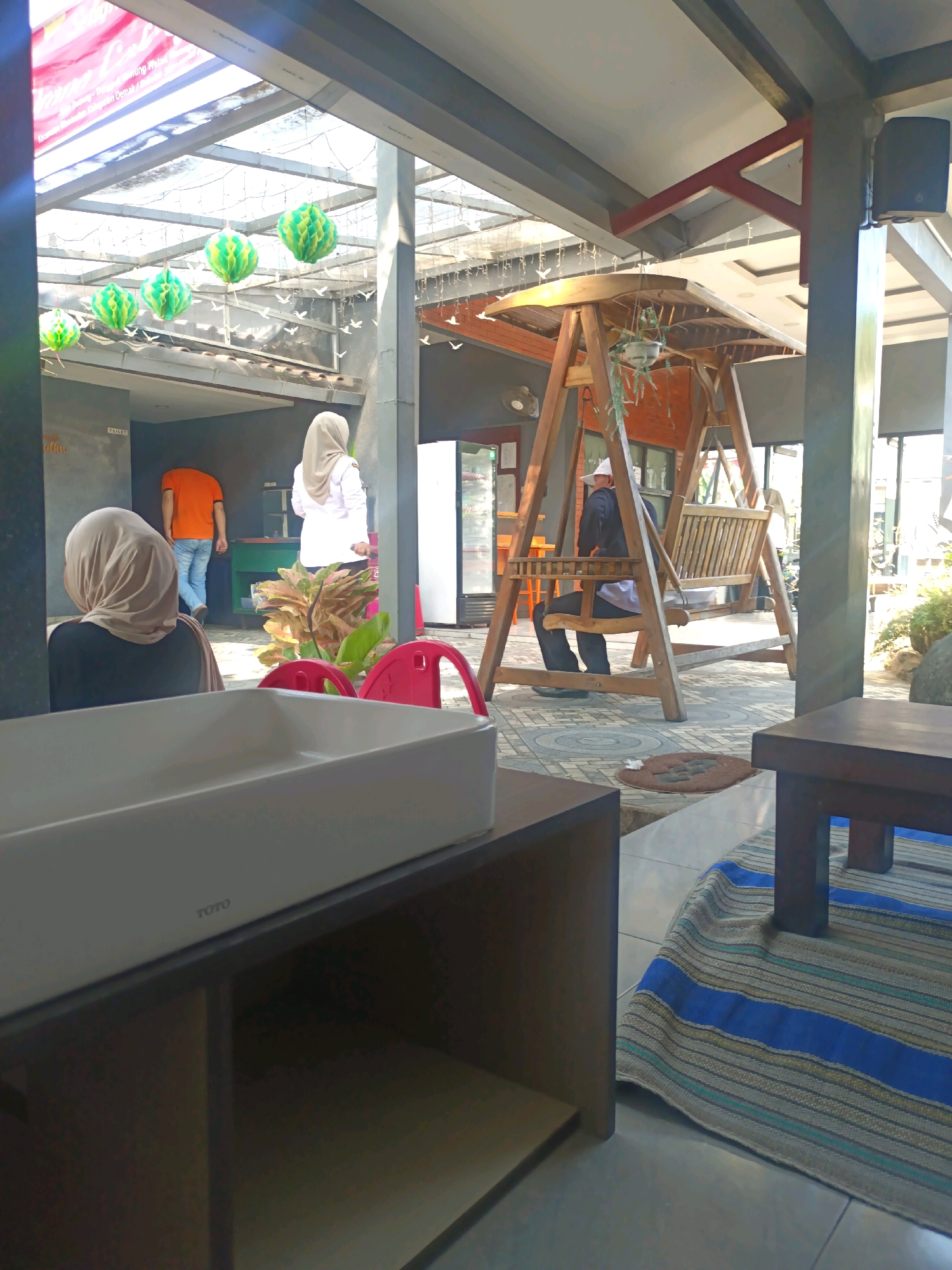 This image depicts an outdoor seating area at a restaurant, likely in Kerangkulon, Indonesia, with a focus on a modern, minimalist design. In the foreground, a white TOTO sink is visible on a dark wooden cabinet. The background shows a wooden swing set with a person seated on it, facing away from the camera. Other individuals are also present, including two women wearing hijabs and a man in an orange shirt. The environment includes a covered patio area with a translucent roof, green decorative lanterns hanging from the ceiling, and various plants. The floor is tiled, with a patterned rug visible in the lower right corner. The overall impression is a relaxed, open-air dining space.