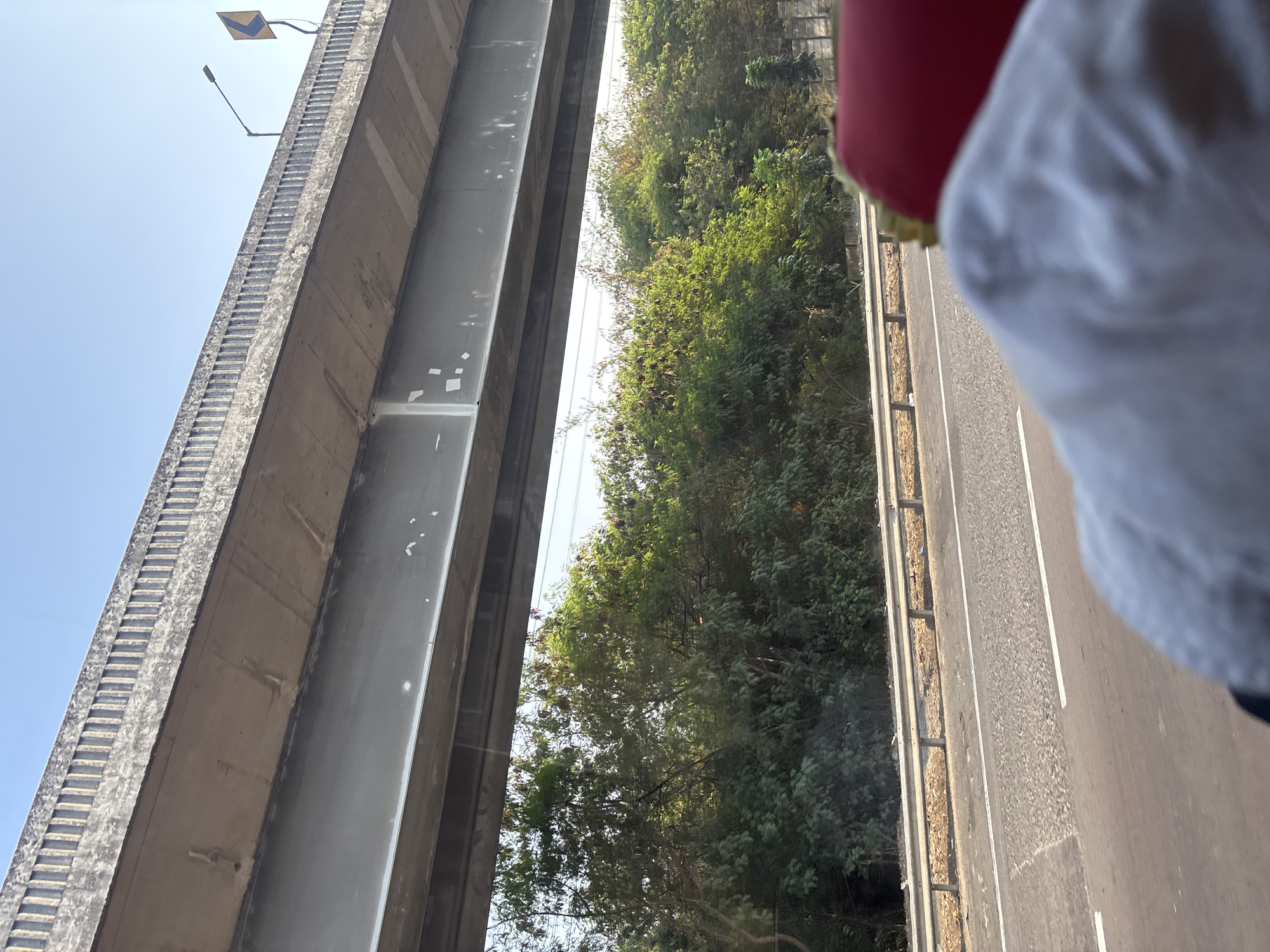 The image shows a view from a vehicle traveling on a highway in Karawang, Indonesia. An elevated highway structure dominates the upper portion of the frame, casting a shadow below. The sky is clear and blue. Below the elevated highway, a dense line of green trees and foliage is visible. A metal guardrail separates the highway from the vegetation. The road surface appears to be asphalt with white lane markings. In the foreground, the blurred image of a person's head wearing a light-colored cloth is visible, suggesting the photo was taken from inside a moving vehicle. The overall scene depicts a typical highway landscape in an urban or semi-urban area.