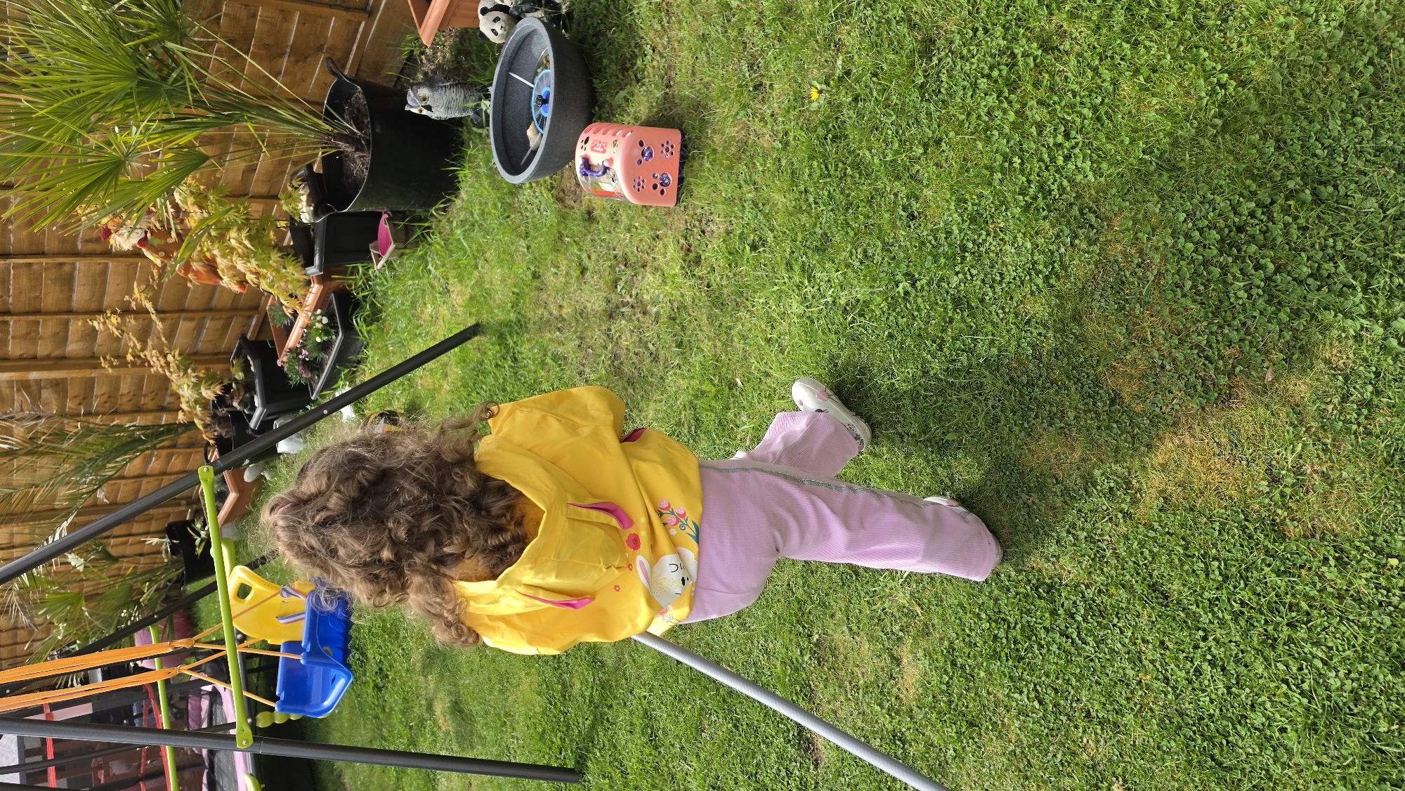 A child with long brown curly hair, wearing a yellow hooded jacket and pink pants, is standing on a green lawn next to a swing set. The location appears to be a garden, with a wooden fence and various potted plants. In the background there are some plant pots, one with pandas on and some other garden items. The lawn consists mostly of short, green grass. The specific location in the city of London, United Kingdom cannot be verified from the given image.