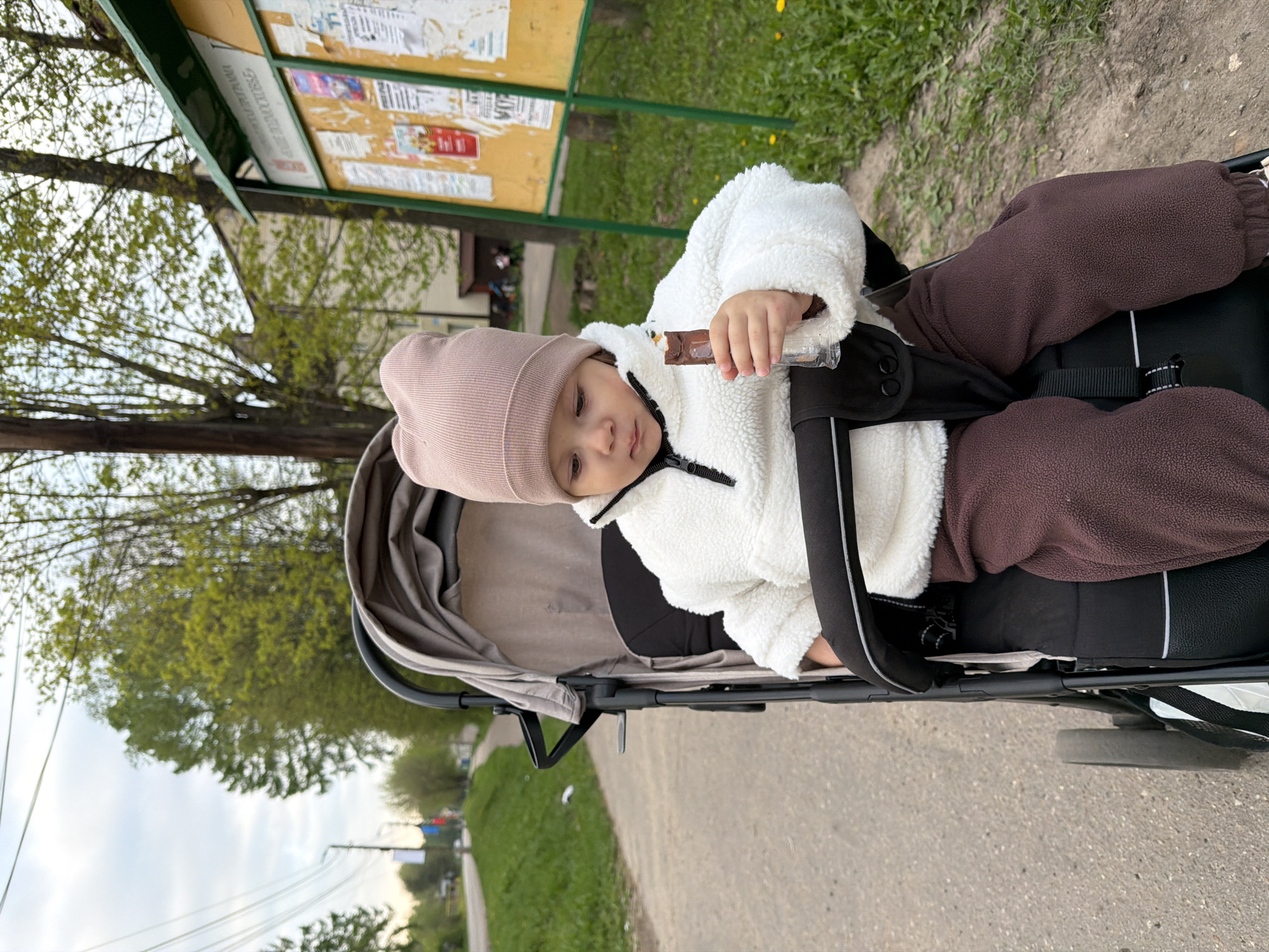 A child, wearing a pink hat and white fleece jacket with brown pants, sits in a black stroller and holds a chocolate bar in their left hand. The stroller is on a gray asphalt road, with green grass visible on either side. In the background, a bulletin board is attached to a pole, displaying various advertisements. Trees are also visible in the background. The location is identified as Reshetnikovo, Russia.