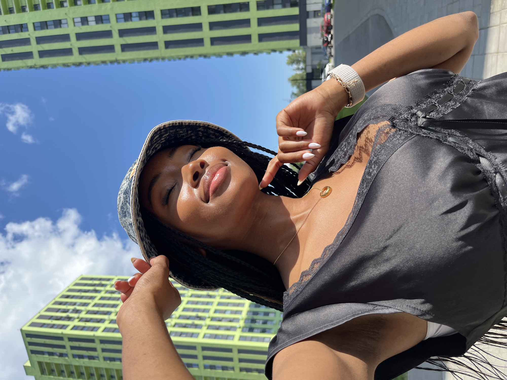 The image is a selfie taken outdoors in front of two identical, tall, lime-green apartment buildings with dark vertical accents. The sky is a clear, bright blue with a few scattered white clouds. A young Black woman, wearing a black slip dress with lace trim, a patterned bucket hat, and a gold pendant necklace, is the primary subject. Her eyes are closed, and she has a gentle smile on her face. Her right hand is raised, touching the brim of her hat, and her fingernails are painted white. A silver and white bracelet is visible on her left wrist. In the background, behind the buildings, there is a parking lot with several cars. The overall setting appears to be a modern urban environment. The location is identified as Kazan, Russia.