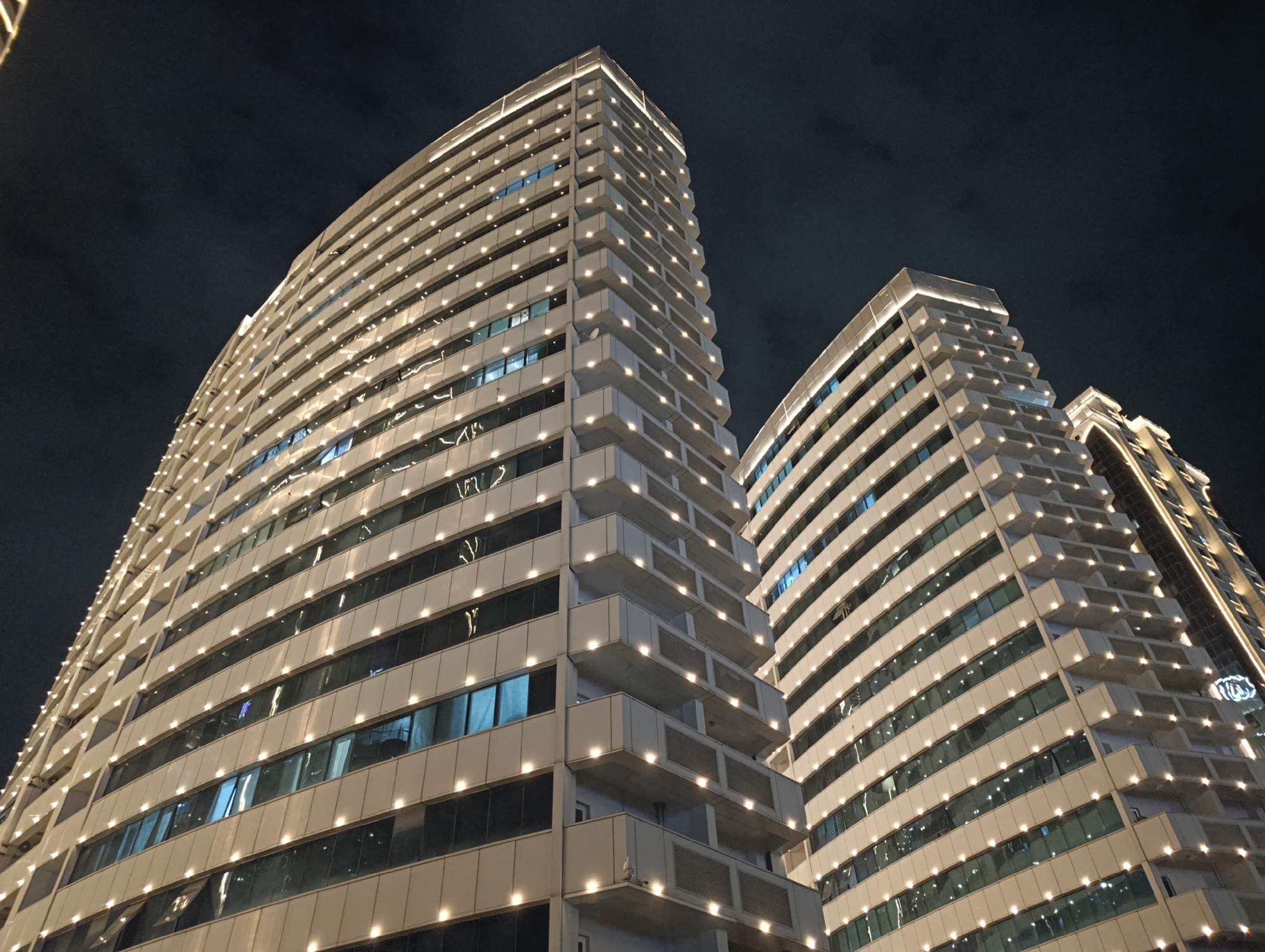 The image shows a low-angle view of two modern skyscrapers at night in Grozny, Russia. The buildings are tall and angular, with a light-colored facade and numerous windows. Each floor is illuminated by small, bright lights positioned along the outer edges of the balconies or facade. The sky above is dark and cloudy. The skyscraper in the foreground is larger and closer, dominating the left side of the frame. Another, slightly shorter skyscraper is visible to its right, set back and partially obscured. The lighting on the buildings creates a grid-like pattern of illumination against the dark sky.