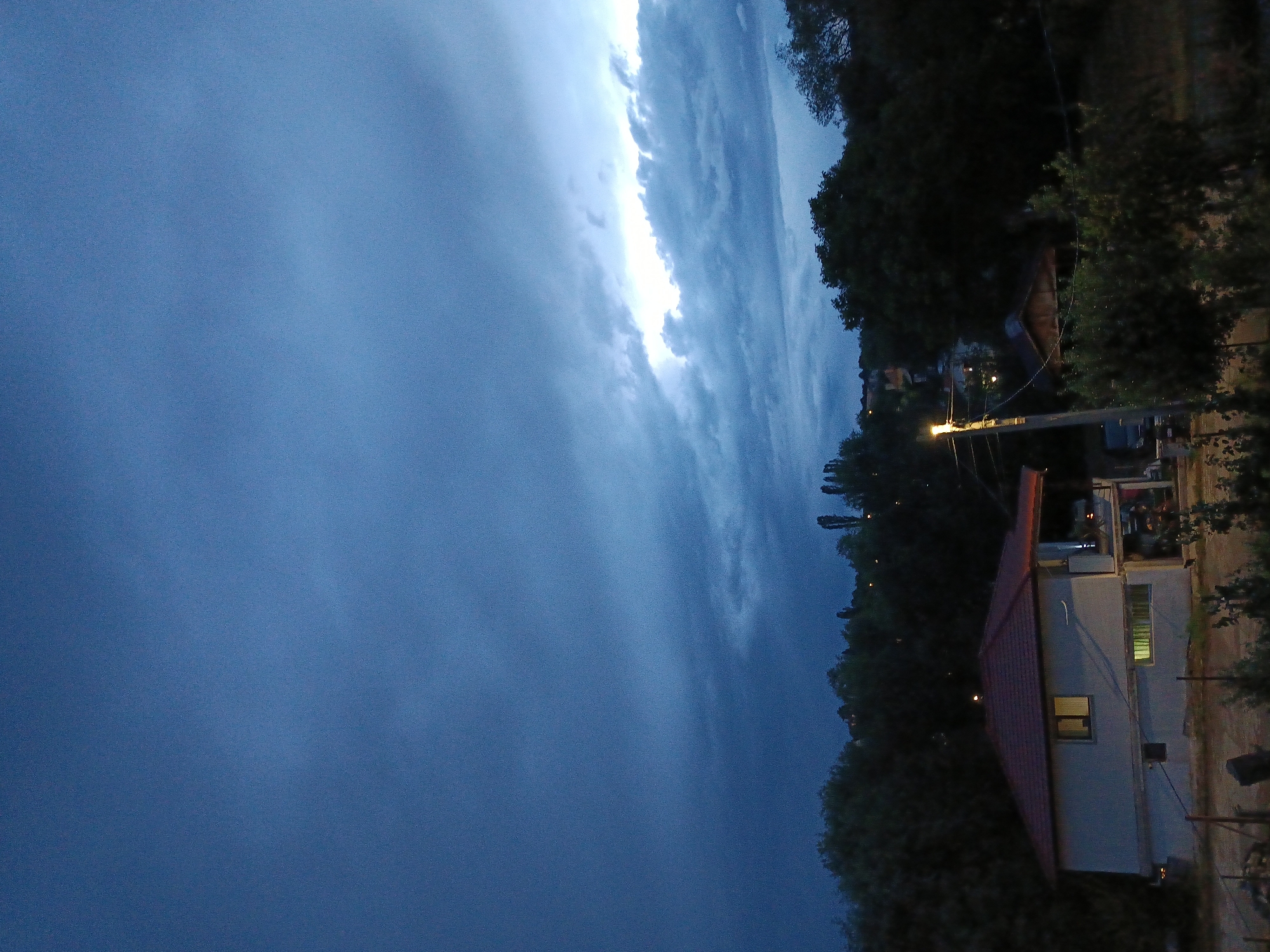 The image depicts a dramatic sky at dusk, with dark, heavy clouds ominously gathering. A bright streak of light cuts through the center of the frame, suggesting a break in the clouds or possibly the edge of a storm. To the right, a residential area is visible, nestled amongst dense trees. A house with a red tiled roof is prominent, its windows illuminated, casting a warm glow against the darkening sky. Power lines and poles are also visible, indicating the presence of infrastructure. The overall scene evokes a sense of anticipation, possibly of an impending storm. The time of day is clearly dusk, with the natural light fading and artificial lights beginning to appear. The weather appears turbulent, with dark clouds dominating the sky. The location cues, combined with the provided context, suggest a rural or semi-rural setting in Kralev Dol, Bulgaria. There is no visible text in the image.