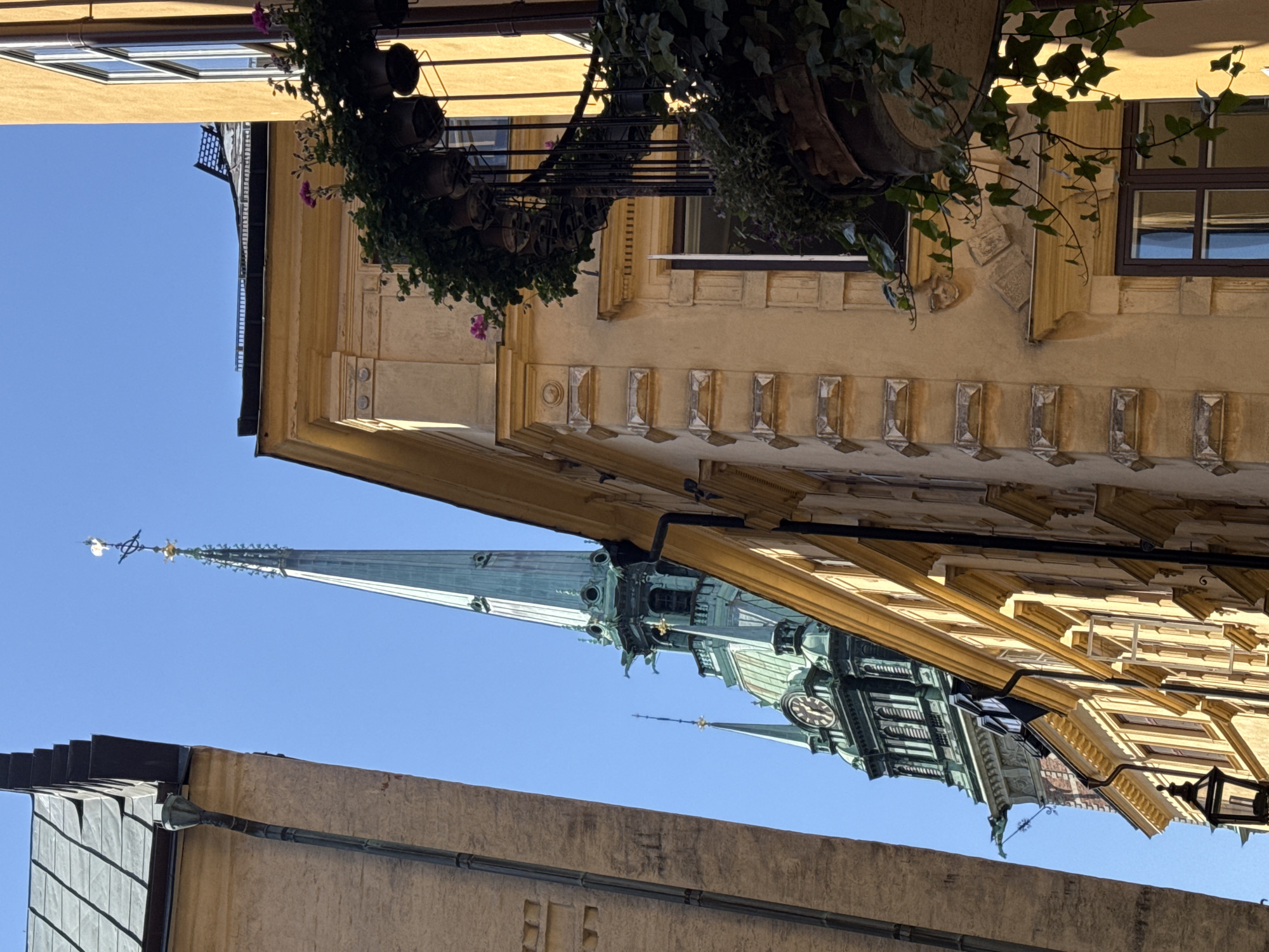 This is a medium shot, eye-level, outdoor, daytime, low-angle photograph taken from a narrow street in Stockholm, Sweden. The primary subjects are the spires of two churches, one taller and more prominent than the other, both featuring green, copper-like roofs and ornate architectural details. A clock face is visible on the taller spire, indicating the time is approximately 3:15. The buildings framing the street are in varying shades of yellow and beige, with visible architectural embellishments, including cornices, decorative carvings, and windows. A balcony adorned with potted plants and hanging ivy is present on the right side of the frame, adding a touch of lushness to the urban landscape. The sky is a clear, bright blue. There are no people visible in the frame. The overall scene evokes a sense of historic European architecture and a sunny day.