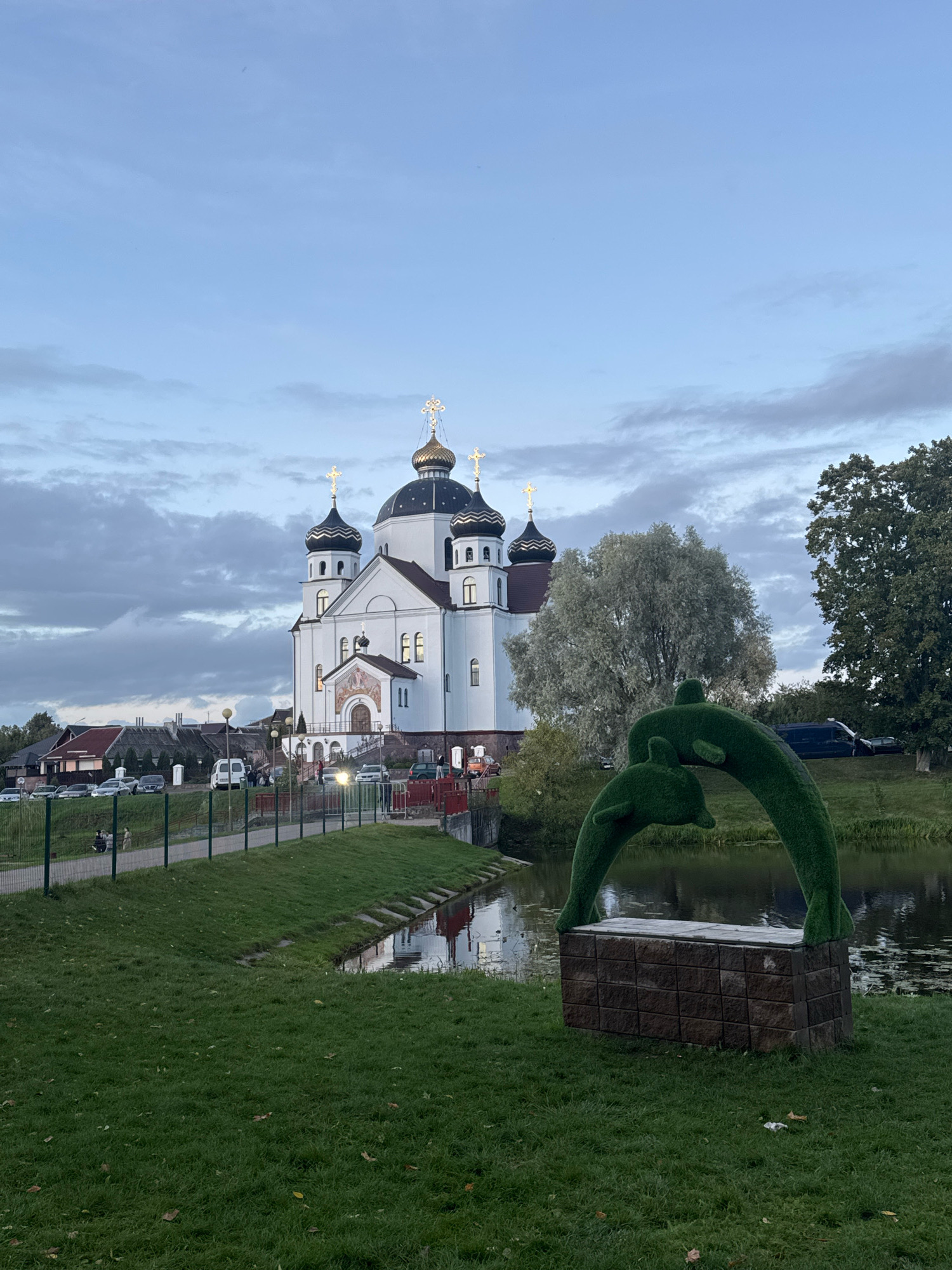 The image depicts the St. Sergius of Radonezh Church in Smarhon, Belarus, situated on a slight incline. The sky above is overcast with a mix of blue and grey clouds. In the foreground, a meticulously manicured green lawn slopes down towards a body of water, with fallen leaves scattered across the grass. A decorative sculpture of two green dolphins, resembling topiary, is prominently displayed on a stone pedestal near the water's edge. A pathway with a metal fence runs along the side of the lawn, separating it from a parking area where several cars are visible. Beyond the parking area, a cluster of houses with red roofs can be seen. The church itself is a large, white building with multiple dark domes topped with golden crosses, characteristic of Orthodox architecture. The church has several windows and an entrance with a mural above it. A few individuals are present near the church entrance and on the pathway. The overall scene suggests a late afternoon or early evening setting, with ambient lighting reflecting off the water and the church's facade.