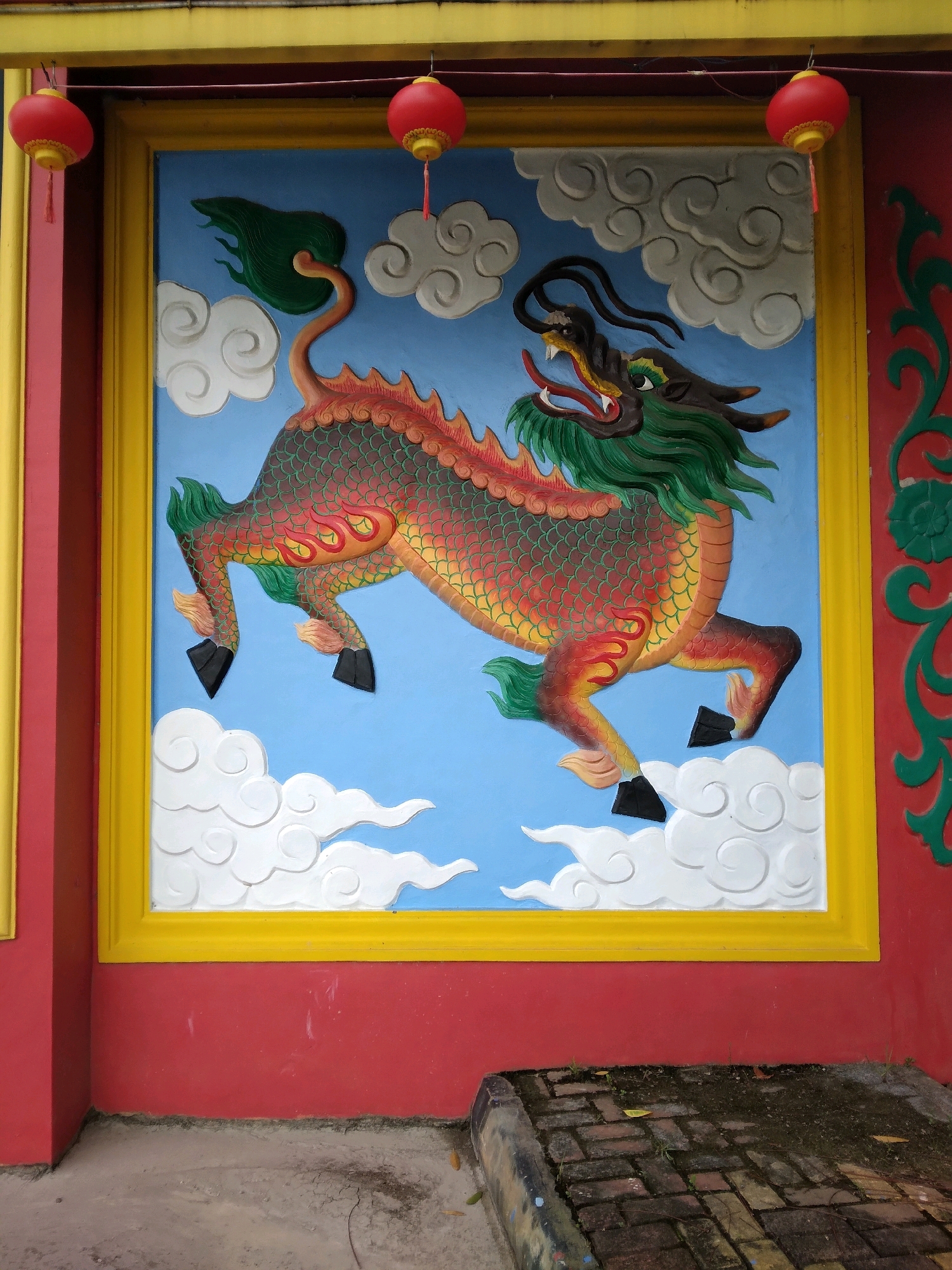 A colorful, three-dimensional mural of a Chinese dragon is mounted on a wall, framed by a yellow border and set against a blue sky with white clouds. The dragon is depicted in motion, with scales of red, orange, and yellow, a green mane, and black hooves. Red lanterns with yellow accents hang from a wire above the mural. The wall itself is painted red, with a yellow border and some green decorative elements on the right side. The base of the wall rests on a textured concrete surface with some vegetation. The scene is outdoors, with part of a brick pathway visible at the bottom right. Based on the architectural style and decorative elements, this mural is likely located at a Chinese temple or cultural center in Batam, Indonesia.