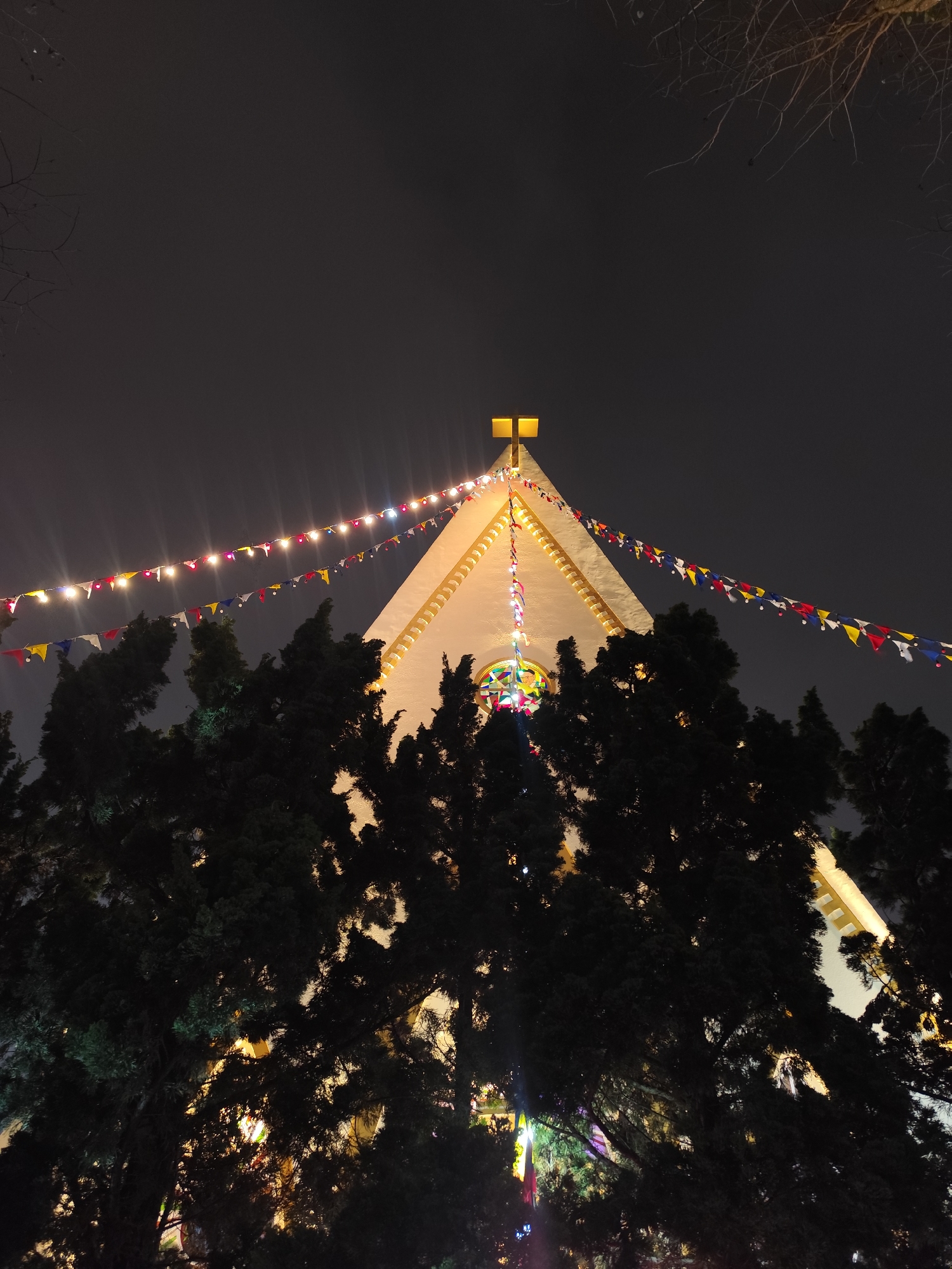 A low-angle shot shows the facade of a white building with a steeple topped by a lit cross, likely a church, at night. The building is adorned with strings of colorful pennant flags and lights. A large, dark mass of trees obscures the lower portion of the building. The sky is a uniform, dark gray, indicating it's nighttime. Based on the style of architecture and the decorations, it is likely located in Da Lat, Vietnam, based on the location mentioned.