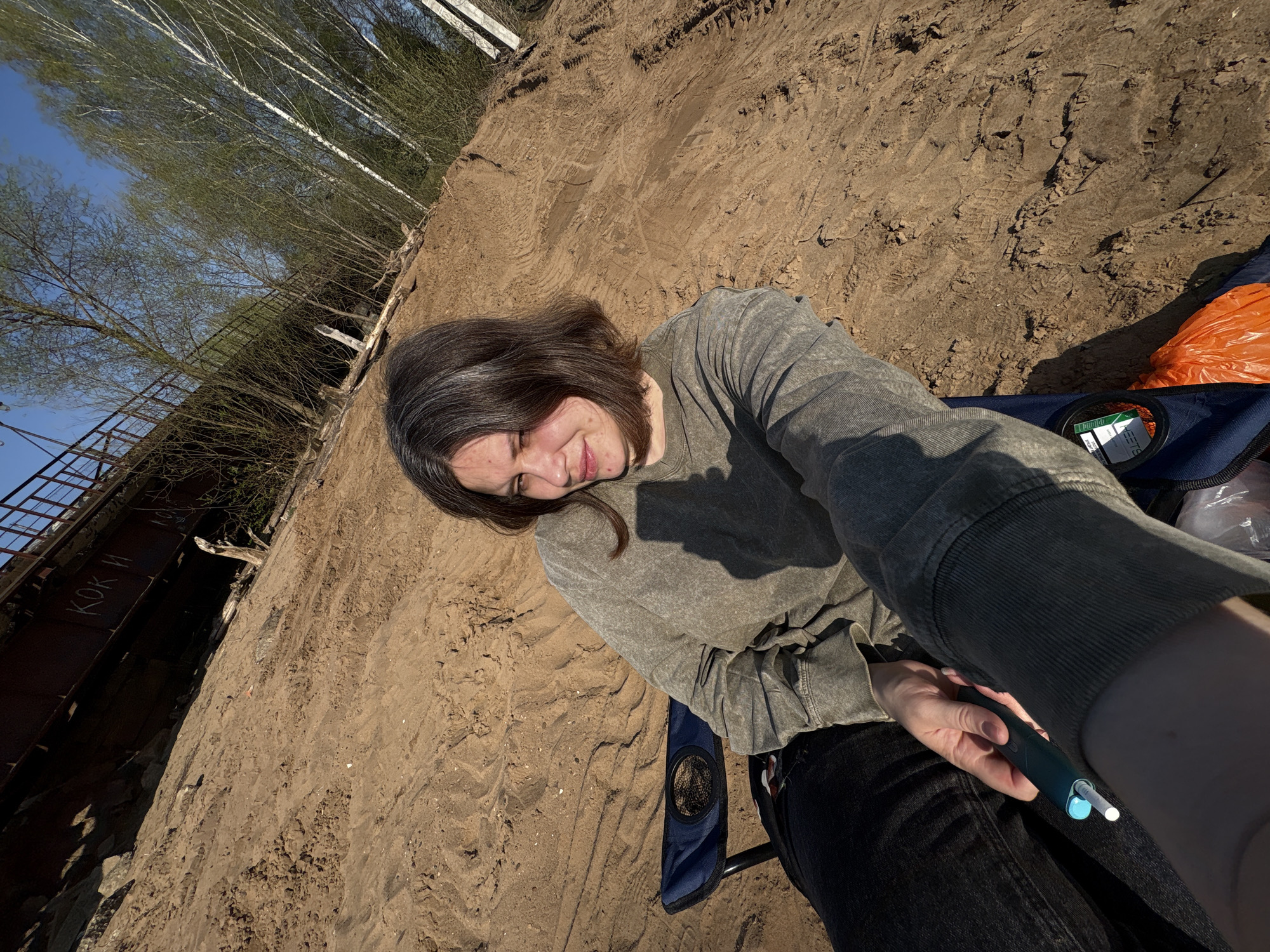 A woman sits on a folding chair on a sandy bank. She is wearing a long-sleeved grey top and black jeans and holds a dark blue electronic device in her right hand. An orange plastic bag is near the chair. The bank slopes upwards to the left, revealing a metal bridge with some Russian text “КОК И”. Trees are visible above the bank, and the sky is blue. This location is likely a bank of the Kama River in Chaikovsky, Russia, given the Russian text on the bridge and typical riverbank landscape.