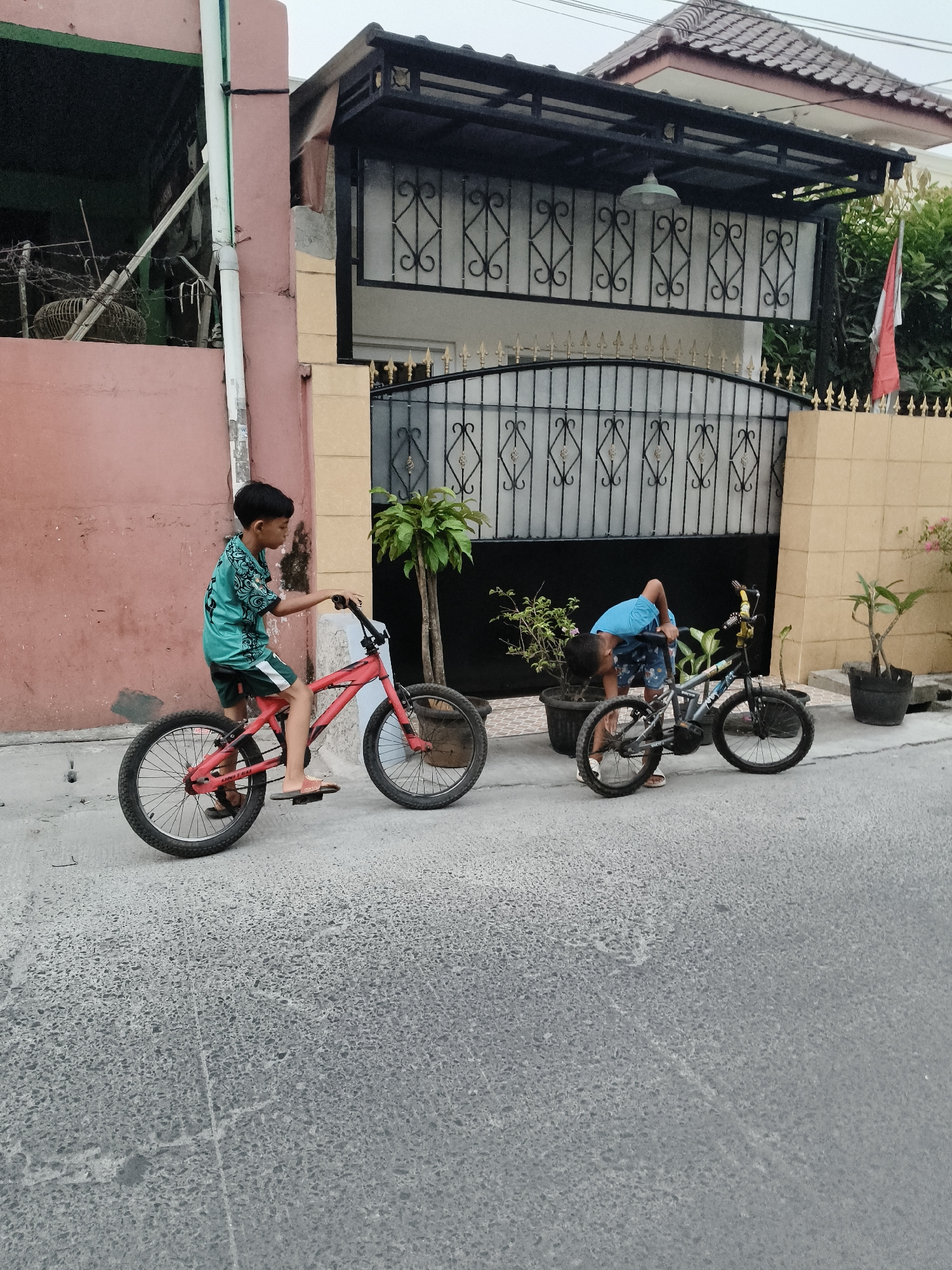 In a residential street in South Tangerang, Indonesia, two young boys are engaged with their bicycles. The first boy, positioned on the left side of the frame, is seated on a red bicycle, appearing to be ready to ride. He wears a teal t-shirt with a patterned design and dark shorts. The second boy, on the right, is bending over his grey bicycle, seemingly making an adjustment or inspecting it. He is dressed in a blue t-shirt and patterned shorts. Both boys are barefoot, wearing flip-flops. The street is paved with asphalt, and a pink wall forms the backdrop on the left, with a house featuring an ornate black gate and tiled roof on the right. Potted plants are situated near the gate. The sky is overcast, suggesting a muted light.