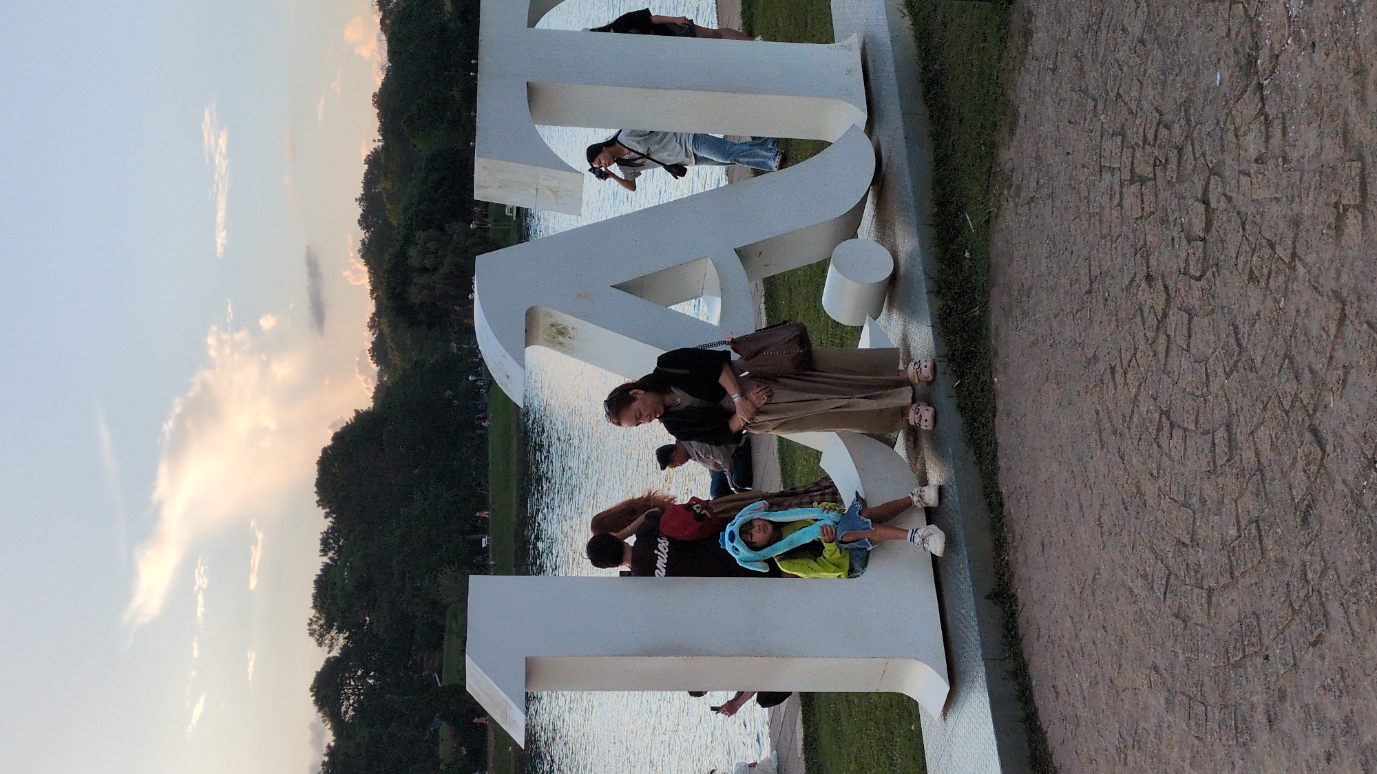 The image captures a serene sunset scene at the "14" sculpture located in Xuan Huong Lake, Da Lat, Vietnam. In the foreground, a young girl wearing a blue Stitch hat and a green long-sleeved shirt sits on the base of the "1" sculpture. Beside her, a woman in a black jacket and beige pants stands, looking downwards. Further back, a couple embraces, with the woman's hair covering her face. Another person is seated in the background. The massive white "14" sculpture dominates the scene, with the numbers crafted from large, white, three-dimensional letters. Behind the sculpture, Xuan Huong Lake stretches out, its surface reflecting the soft hues of the twilight sky. The distant treeline forms a dark silhouette against the horizon, punctuated by scattered clouds that catch the fading sunlight. The ground in front of the sculpture is paved with a textured pattern, and a grassy area separates it from the lake. The overall atmosphere is peaceful, with the natural beauty of Da Lat providing a picturesque backdrop.