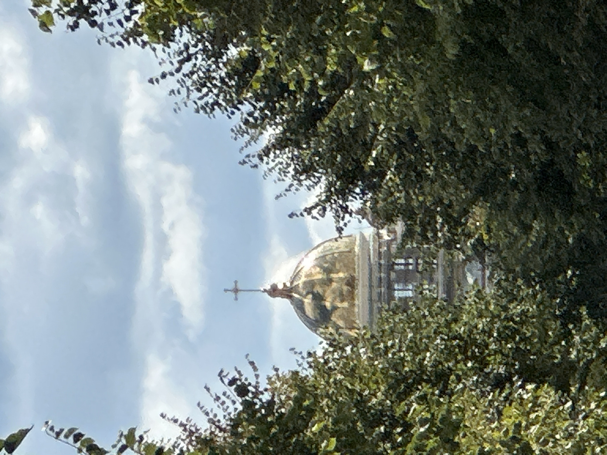 The image shows a view of a domed building with a cross on top, framed by green trees. The building appears to be made of stone and has a golden dome. The sky is cloudy. The provided crops show closer details of the dome, the cross, and the surrounding foliage. There are no people or actions visible in the image. The location is identified as the city of Podolsk, Russia, implying the domed building is a church or a similar religious structure within that city.