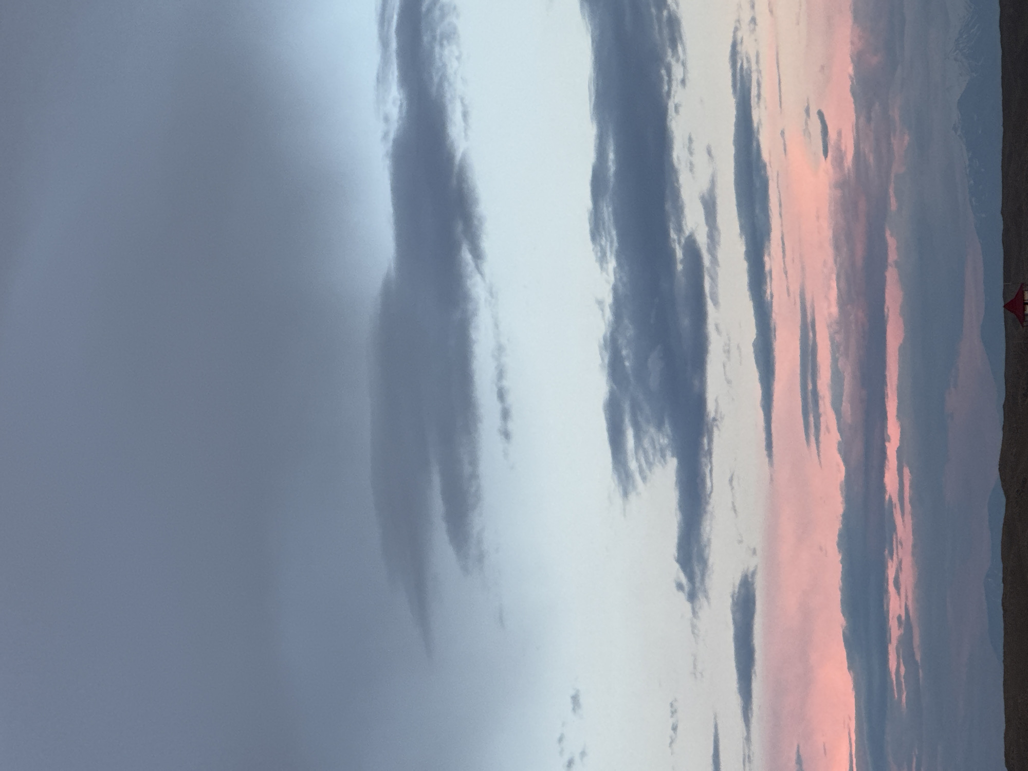 The image shows a landscape at twilight or sunrise, likely in a rural area. Predominantly, the sky is filled with clouds of varying shades of gray, white, and pink, suggesting the presence of a colorful sunset or sunrise. On the right side of the image, a small patch of land is visible, with some scrub vegetation. In the distance, there's a small red object that could be a building or a vehicle. The overall setting seems to be an open space with a dramatic sky. There is no way to know if this scene took place in Anipemza, Armenia without further context.