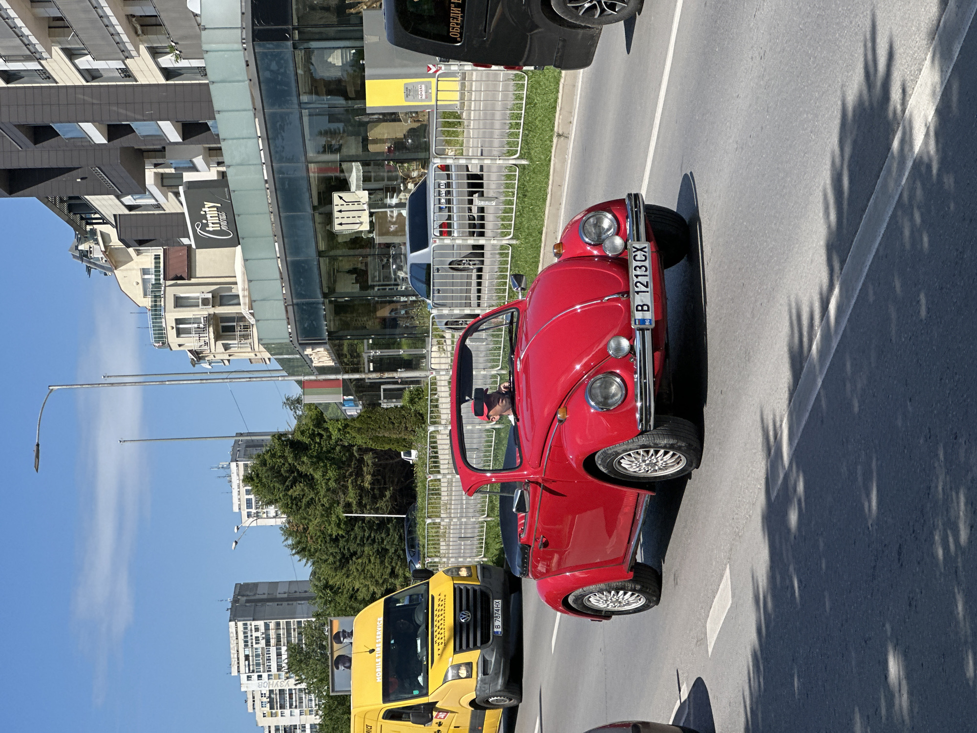 The image captures a street scene in Varna, Bulgaria. A red Volkswagen Beetle cabriolet, with license plate "B 1213 CX", is in the foreground on a grey asphalt road. To its left, a yellow "MOBILE TIRE SERVICE" van, with license plate "B 7874 HX", is also parked. A man can be seen driving the red Beetle. The road is lined with a white line marking the lane. Shadows of trees are cast on the road's surface. In the background, there is a modern building with the inscription "Trinity" on it, and several other buildings. Green trees and a blue sky with light clouds are also visible. Fences are present alongside the road.