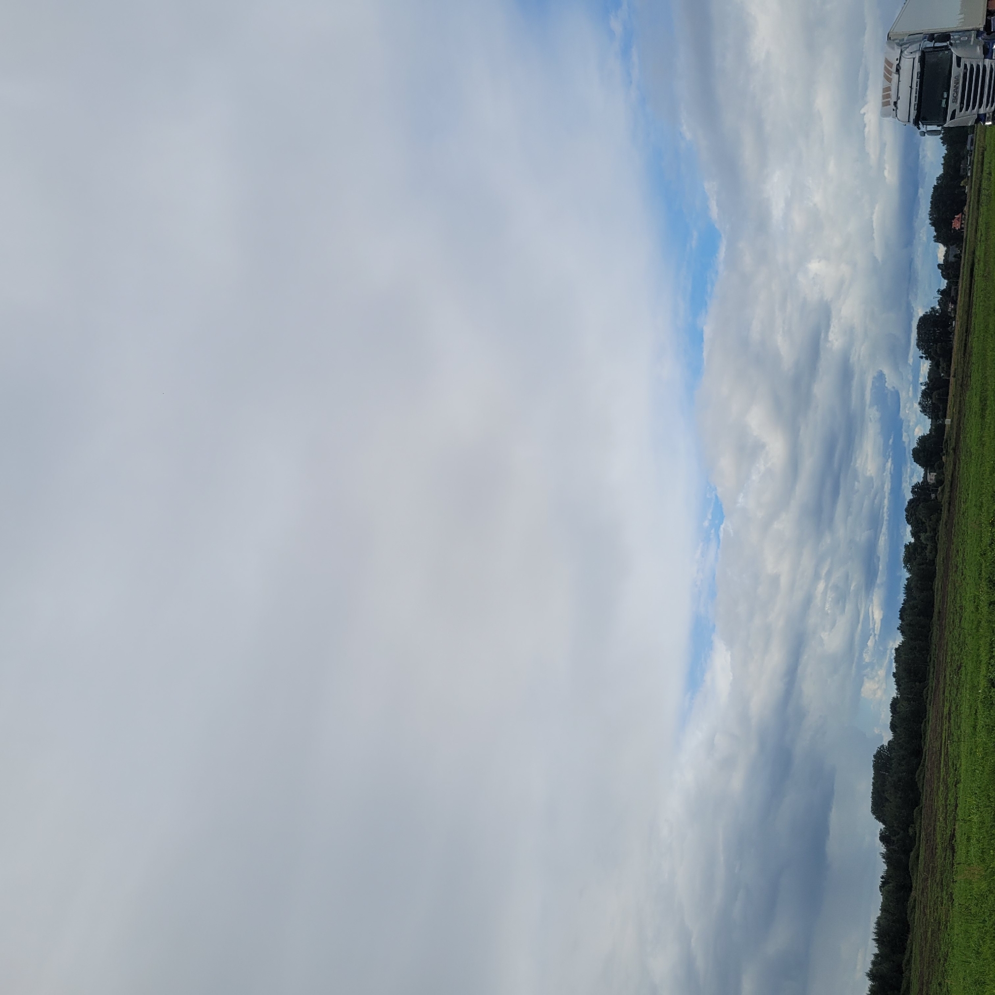 The image depicts a wide, overcast sky with patches of blue visible through the clouds. In the foreground, a field of green grass extends to a line of dark green trees. To the right of the frame, a white semi-trailer truck with "SCANIA" written on its grille is partially visible, suggesting it is on or near a road adjacent to the field. The overall scene is rural, consistent with the location of Jaunmārupe, Latvia.