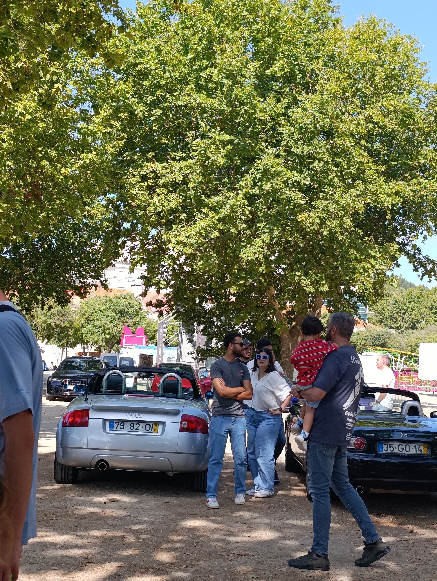 In Setúbal, Portugal, under the shade of a large leafy tree, a group of people are gathered around several parked cars. In the foreground, a silver Audi TT convertible is parked. Its license plate reads "79-82-OI". To the right of the Audi, a black Mazda convertible is visible, with its license plate showing "35-GO-14".
Several individuals are in view. A man in a dark blue t-shirt, carrying a young child in a red and white striped shirt, is walking away from the camera. The child is looking towards the front. To the left of this man, a woman wearing a white top and blue jeans stands with her arms crossed, looking towards the Audi. Next to her, a man in a grey t-shirt and blue jeans is also looking towards the car, with his arms crossed. Behind them, another person is partially visible. Further back, to the right, another man in a white t-shirt is looking towards the scene.
The ground is covered in gravel and fallen leaves, suggesting an outdoor, possibly park-like setting. In the background, buildings and more trees can be seen, along with some temporary structures and event equipment, hinting at a possible outdoor event or gathering. The sky is clear and blue.