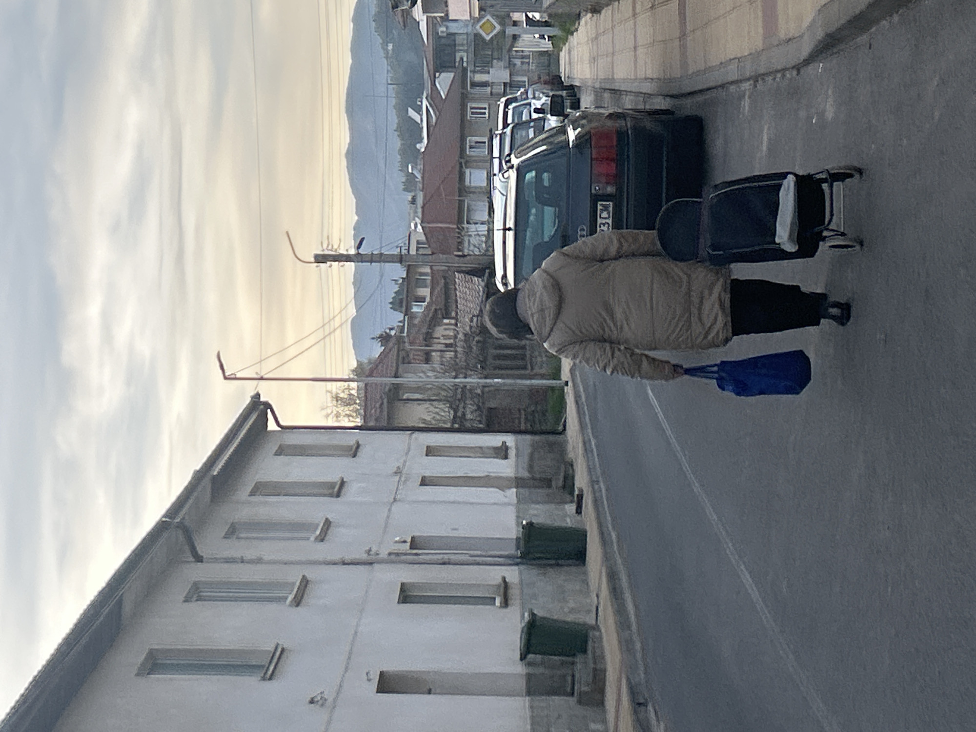 A person is walking down a street in what appears to be Zlatograd, Bulgaria, pulling a black roller suitcase. The individual is wearing a light brown jacket and black pants, and carrying a blue bag in their hand. The street runs past a light-colored building with multiple rectangular windows and green trash receptacles along the base. Behind the person and the car parked on the street are other buildings, including a vehicle with license plate, and a mountain range is visible in the background under a cloudy sky.