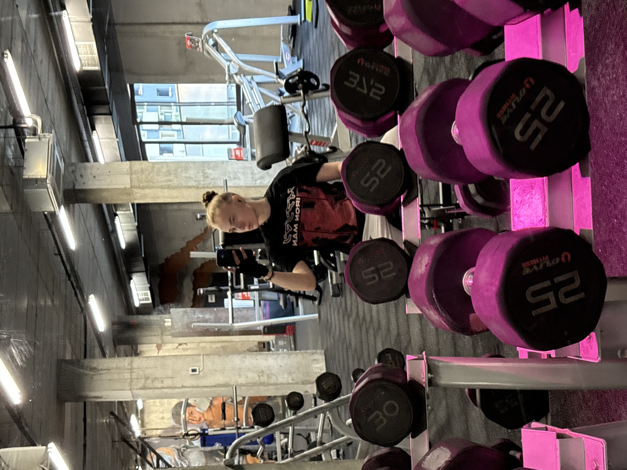 The image shows a gym interior, with a young adult holding a smartphone in the middle of the frame. In the foreground, a rack of purple dumbbells of varying weights is visible. Behind the individual, workout machines and other gym equipment are present. The building has concrete columns, exposed ductwork, and large windows with city views. While the exact location in Sofia, Bulgaria is unclear from the image alone, the architecture and gym equipment are indicative of a modern fitness facility within an urban environment.
