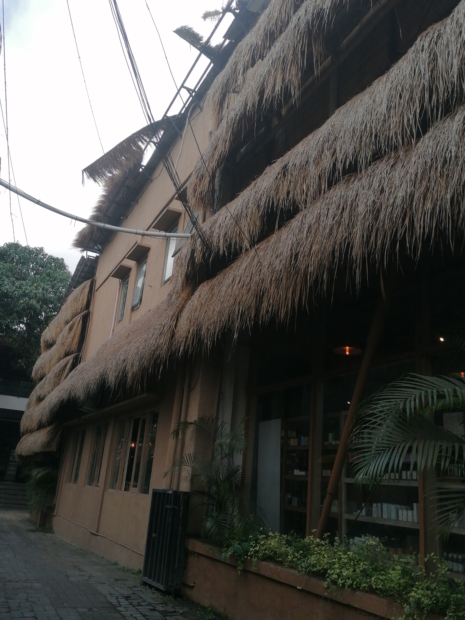 The image shows a three-story building with thatched roofs. The building is tan colored with several windows. There are trees and plants around the building, and the sky is visible above. The building appears to be a shop or restaurant, as there are shelves with items visible through the windows. The area looks like a paved walkway in front of the building. The location is in the city of Ubud District, Indonesia.