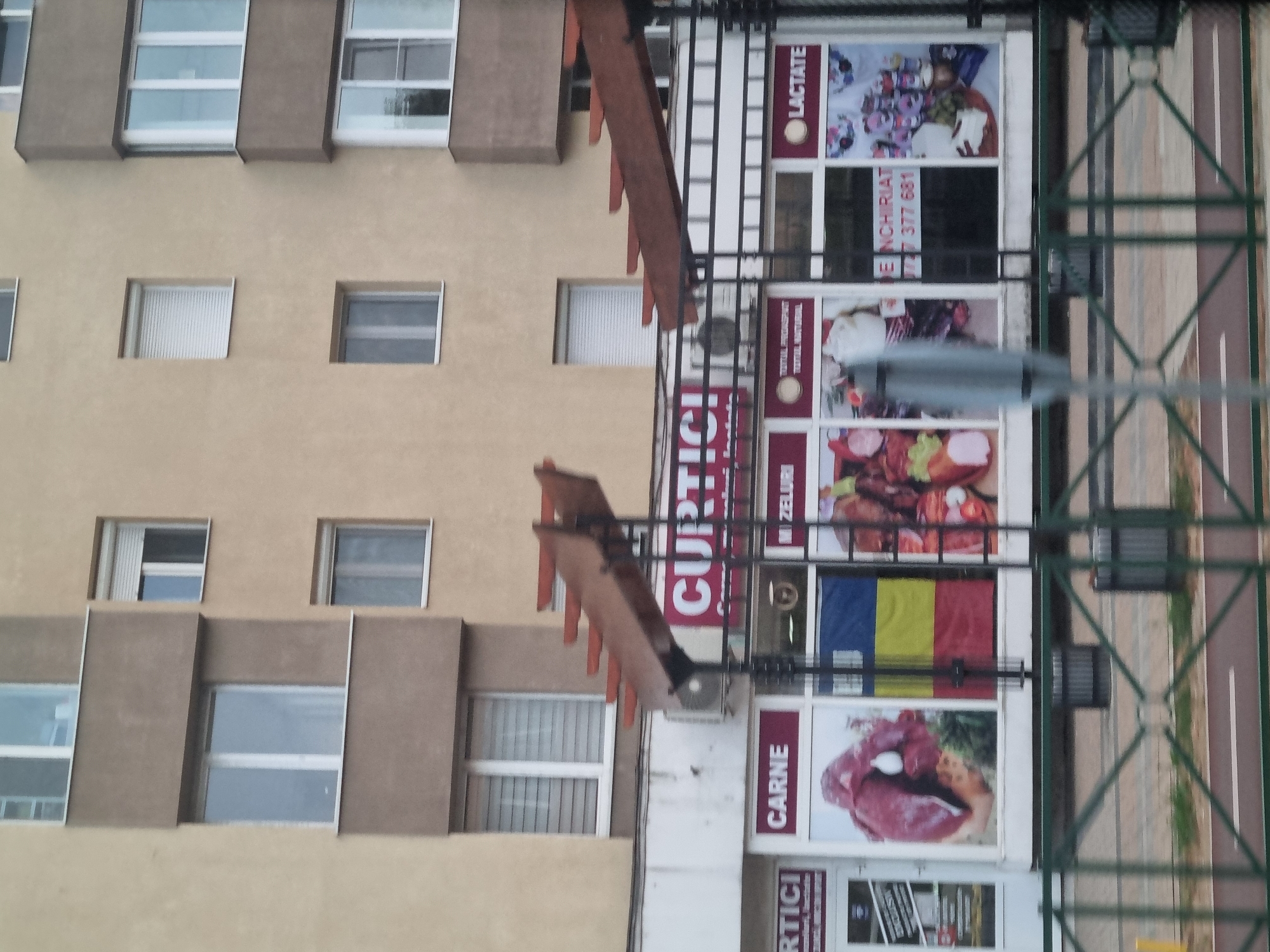The image shows a storefront in Salonta, Romania, with the name "CURTICI" prominently displayed in red lettering above the entrance. The store appears to be a grocery or butcher shop, with sections labeled "CARNE" (meat) and "LACTATE" (dairy products) visible on the storefront windows. Various images of food products, including raw meat, cheese, and jars of preserved goods, are displayed on the windows. A Romanian flag is also visible. The building has a light yellow facade and several windows with white frames. A metal fence runs across the foreground of the image.
