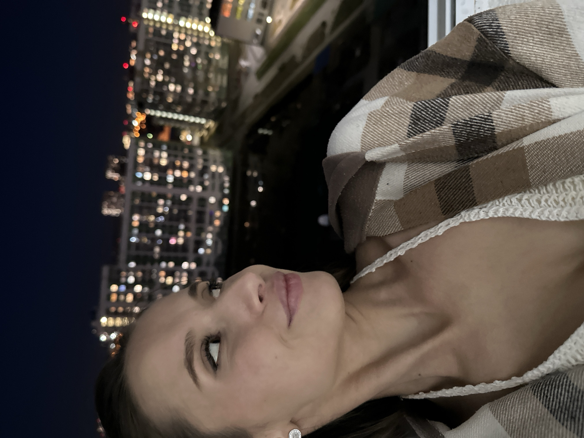 A sideways shot depicts a woman wearing a white crocheted top and a brown and white plaid flannel shirt, seemingly on a balcony. Behind her, blurred lights suggest a cityscape at night, specifically skyscrapers, possibly Yekaterinburg, Russia. The woman is looking upwards, with a relaxed expression. Her right earring is visible.
