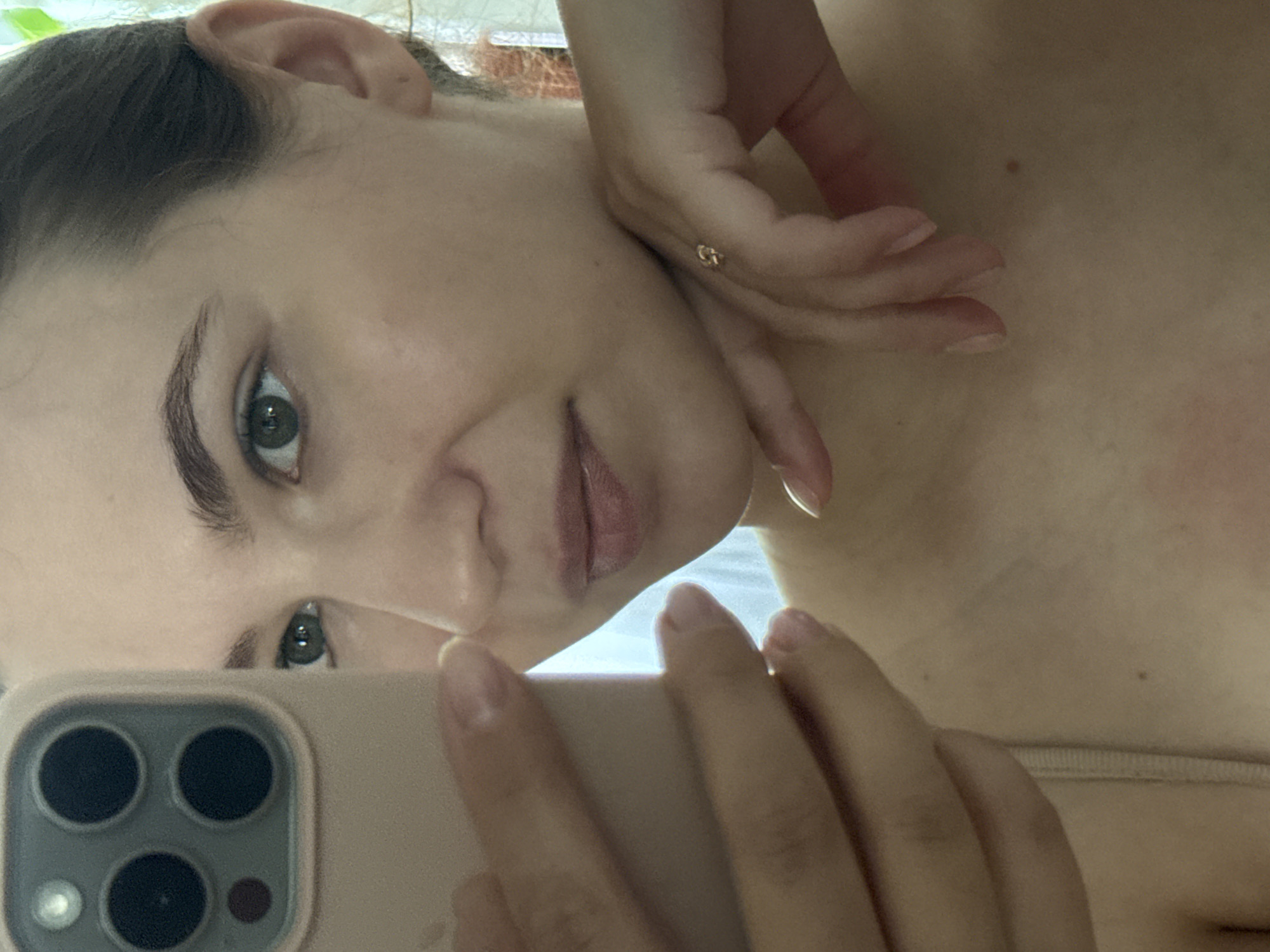 A close-up, waist-up selfie shows a young woman holding a smartphone in her left hand. The phone's camera module is visible on the left side of the image. The woman has light skin, dark eyebrows, and green eyes. Her right hand is resting on her chin, and a small gold ring is visible on her right ring finger. She is wearing a light-colored bra strap. The background is out of focus, showing a blurry, bright light source, possibly a window. The image is a personal photograph, likely taken indoors, and provides no information regarding its location in Chaikovsky, Russia.