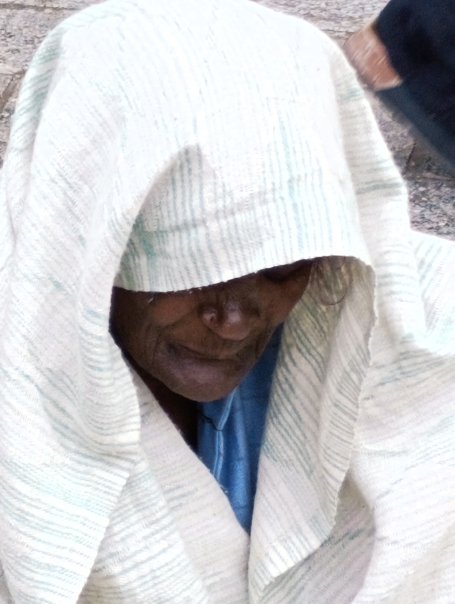 A close-up, vertical, outdoor, eye-level shot focuses on the face of an elderly person, partially obscured by a white, striped cloth. The person, with dark skin and visible wrinkles, is looking slightly downwards. A glimpse of a blue garment is visible beneath the white covering. The background shows a textured, possibly stone or pavement surface and a dark blurred area, suggesting an urban environment. The lighting is diffused, casting soft shadows.