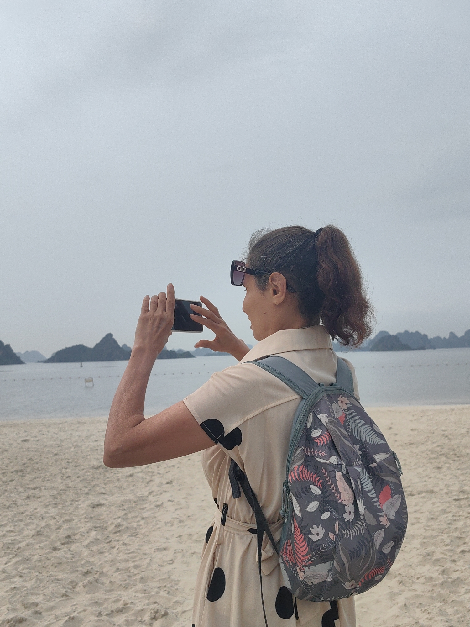 A woman stands on a sandy beach holding a smartphone to take a picture of the scenic view of Ha Long Bay, Vietnam. She is facing away from the camera with her back to the viewer. She has brown hair in a ponytail and is wearing sunglasses. She has a backpack with a floral pattern. She is wearing a light-colored, polka-dot dress. The view beyond her shows water and various karst limestone formations characteristic of Ha Long Bay. The sky is overcast and grey.