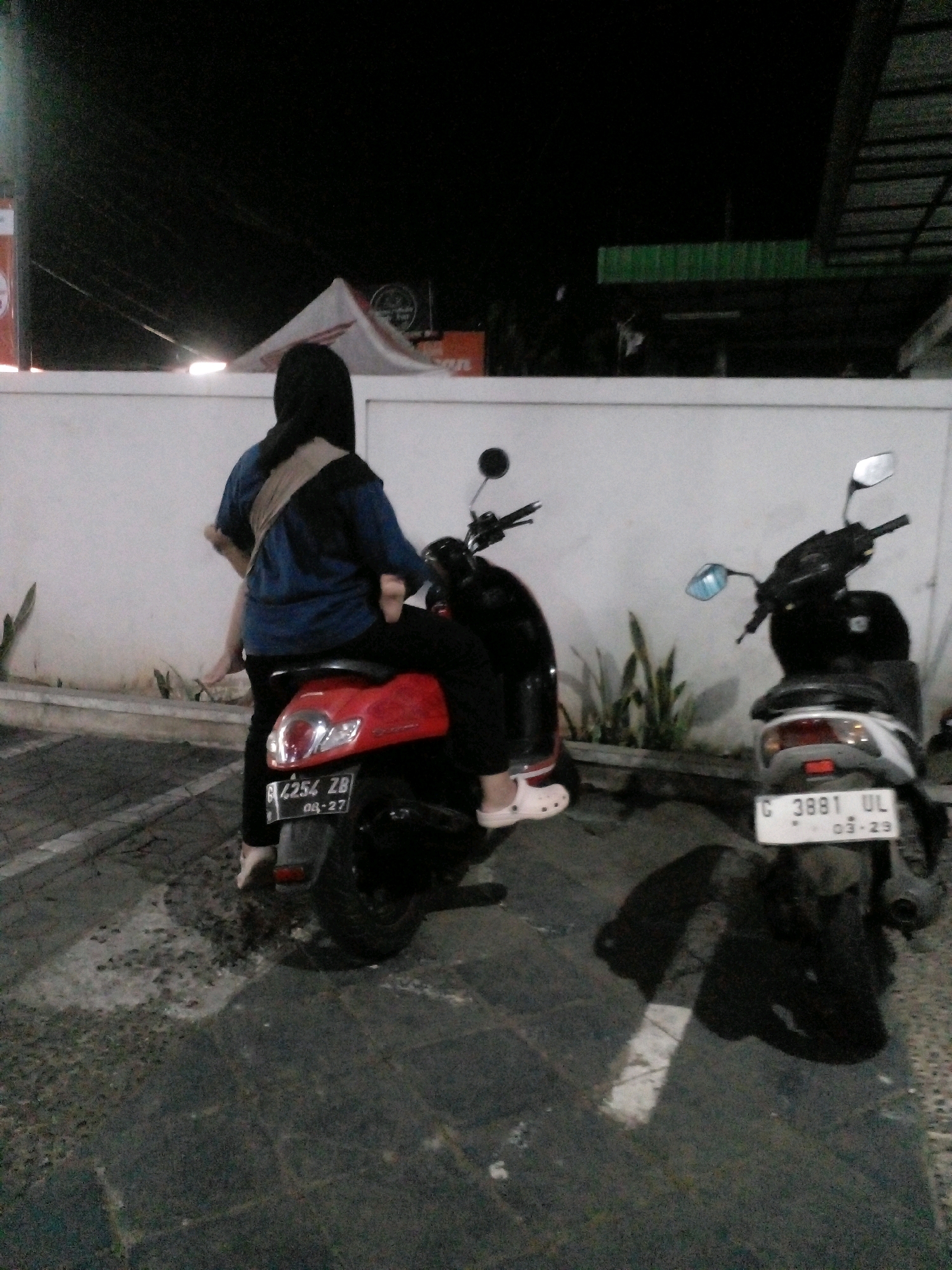 In Banyuputih, Indonesia, at night, a person wearing a black hijab and a blue shirt is sitting on a red scooter. Their legs are crossed, and they are facing away from the camera. Another scooter, white and black, is parked to the right, with its license plate visible. The scooters are parked on a dark, textured surface, possibly pavement or asphalt. In the background, there is a white wall and some greenery. The scene appears to be outdoors, perhaps in a parking lot or on a street.