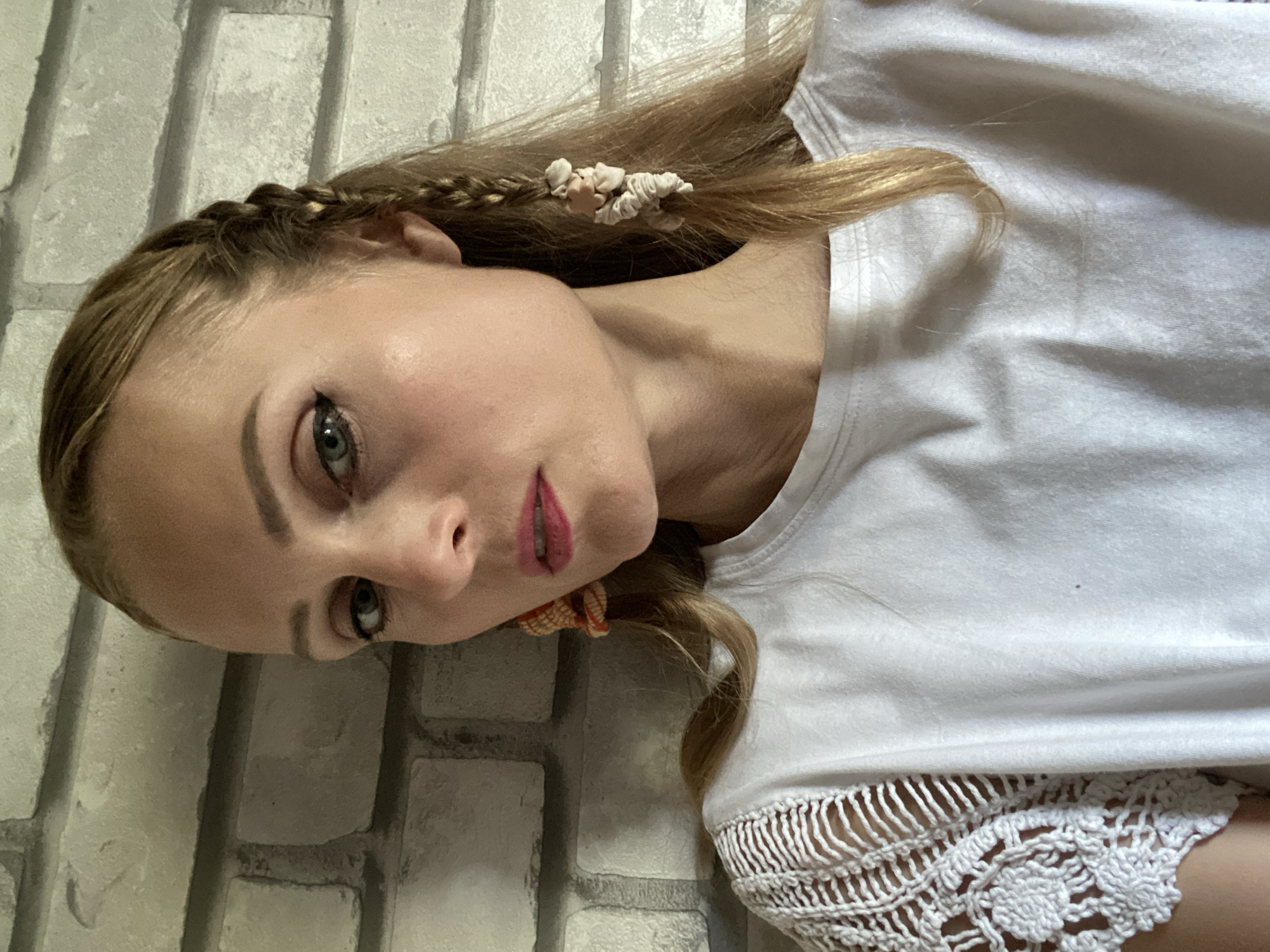 A medium shot captures a fair-skinned woman with blonde hair, styled in a braid with decorative elements, leaning against a brick wall in an indoor setting. She wears a white t-shirt with intricate crochet detailing on the sleeves. Her makeup is subtly applied, with dark eyeliner and pink lipstick. The lighting is soft, casting gentle shadows on her face and clothing. The background consists of a light-colored brick wall. The location is identified as Міханавіцкі сельскі Савет, Belarus, implying the photograph was taken in this region.