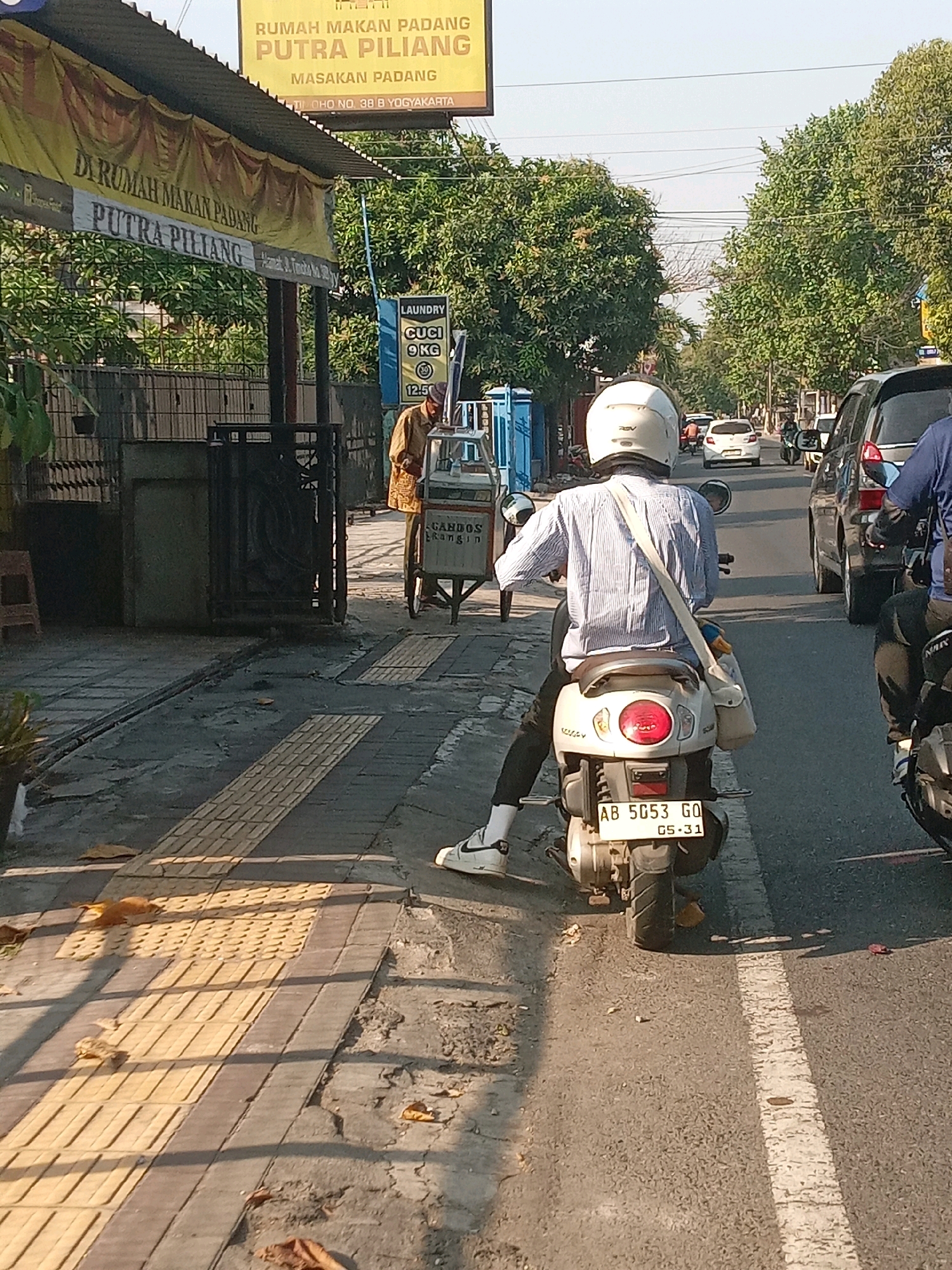 In Yogyakarta, Indonesia, a street scene unfolds under a clear sky. A person rides a white scooter with a license plate reading "AB 5053 GQ 05.31". They are wearing a blue and white striped shirt, black pants, white socks, and white sneakers. A beige bag is slung across their body. The scooter has a red taillight and is seen from behind, with a portion of its rear wheel visible.
To the left of the scooter, a yellow banner with the text "RUMAH MAKAN PADANG PUTRA PILIANG" and "MASAKAN PADANG" is prominently displayed, indicating a Padang restaurant. Below this, another banner reads "DI RUMAH MAKAN PADANG PUTRA PILIANG". Adjacent to the banners is a fence. Further to the left, a person in a brown shirt stands near a cart with wheels. This cart appears to be a food or drink vendor, with text on it that includes "GANDOS" and "IRANGIN". Another cart nearby has a "LAUNDRY" sign with details about "CUCI 9 KG".
The environment features a paved sidewalk with tactile paving for the visually impaired. Fallen leaves are scattered on the ground. A street is visible with several cars and motorcycles on it, suggesting moderate traffic. Trees with green foliage line the street, and power lines are strung across the sky. The overall impression is a bustling urban street in Yogyakarta.