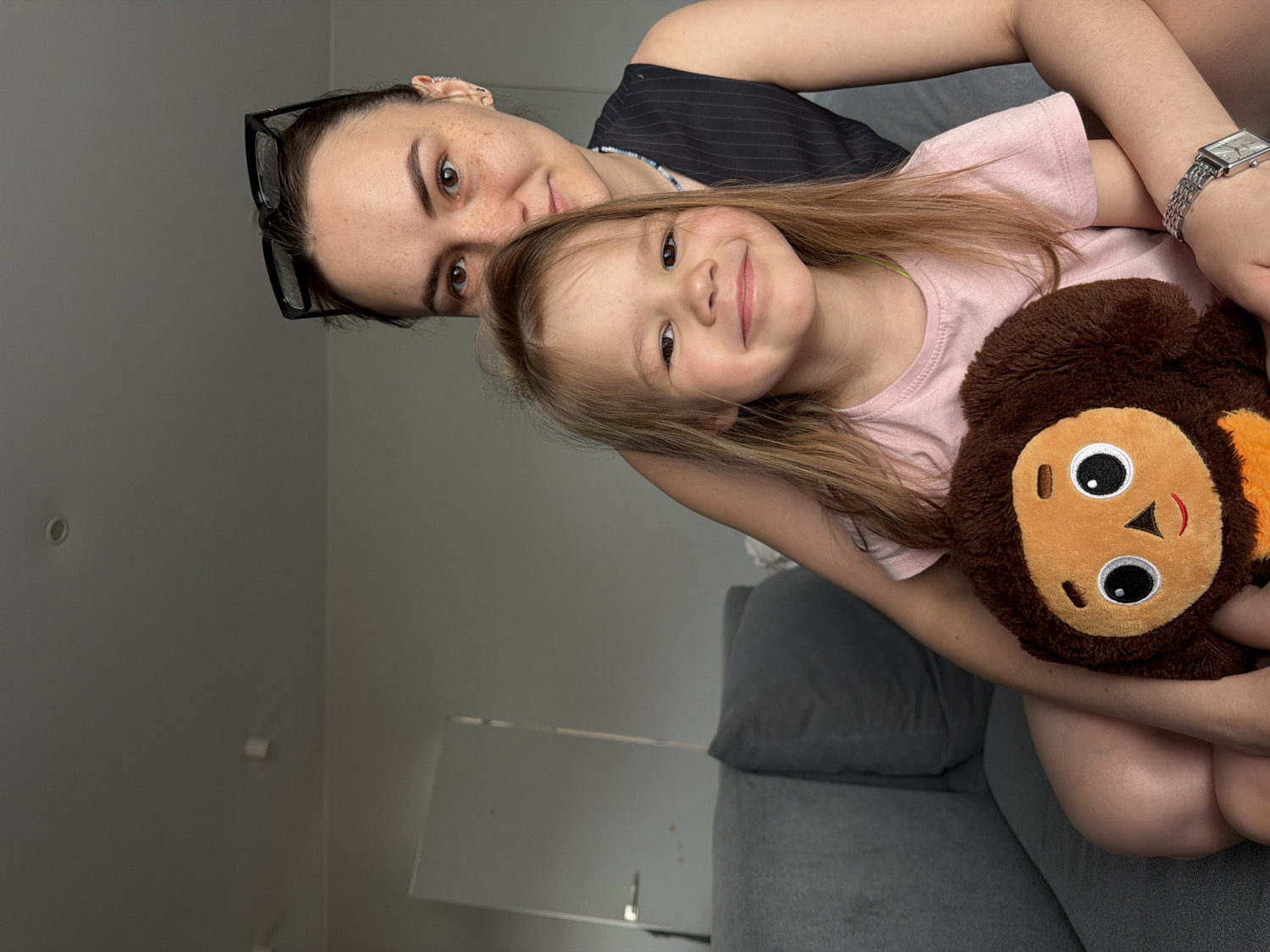 An indoor portrait captures a woman and a young child, likely mother and daughter, in an affectionate pose. The woman, positioned slightly behind and to the left, has dark hair, glasses perched on her head, and a scattering of freckles. She wears a dark pinstriped top and a silver-toned wristwatch on her left wrist. She looks towards the viewer with a gentle smile.
In front of her, the young child, with light brown hair and a bright pink t-shirt, beams a wide, joyful smile directly at the camera. The child is holding a brown plush toy, possibly a monkey, with a lighter face, large eyes, and an orange/red smile. The woman's arm is wrapped around the child and the plush toy.
The background is simple, featuring a plain light grey wall in the upper left with a recessed ceiling light fixture visible. A grey sofa provides a soft backdrop in the lower and right portions of the image. The scene suggests a warm, happy moment shared between the two individuals in a casual home environment, possibly in Chaikovsky, Russia. No visible text is present.