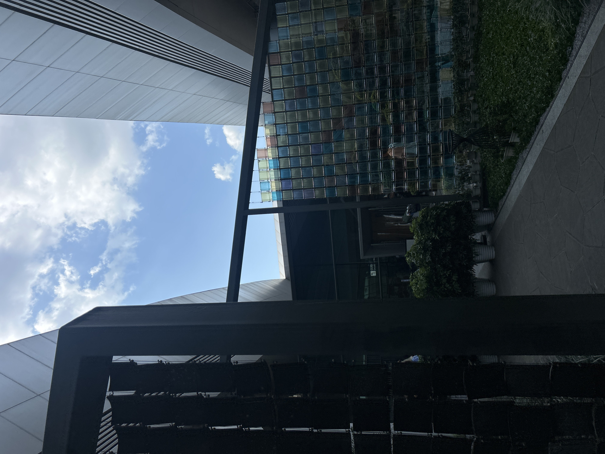The image depicts an outdoor courtyard area adjacent to modern architectural structures. In the foreground on the left, a dark, perforated metal screen forms a partial enclosure. To the right of this screen is a pathway paved with hexagonal and rectangular stones. Beyond the pathway, a lush green lawn is bordered by rows of potted plants. A prominent feature in the middle ground is a wall constructed of translucent glass blocks, displaying a gradient of colors from blue to pink and yellow. Behind the glass block wall, more greenery and architectural elements are visible. In the background, two large buildings with metallic, angled facades dominate the scene, reaching towards a bright blue sky with scattered white clouds. The overall impression is a contemporary urban environment integrating green spaces and modern design.