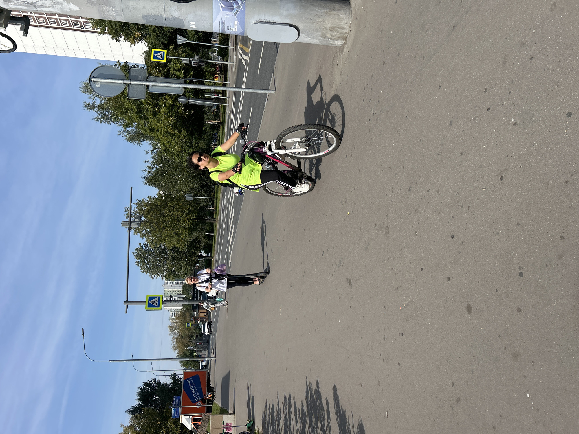 The image shows a paved road in Moscow, Russia, on a sunny day with a clear blue sky. A woman on a bicycle is riding on the right side of the road, wearing a bright yellow top and black leggings. She has sunglasses on and a backpack. To her left, a woman on an electric scooter is also riding on the road, wearing a white top and black leggings, with a pink bag on her scooter. In the background, there are trees, buildings, and road signs, including pedestrian crossing signs. A truck with an orange advertisement is parked on the left side of the road. The scene depicts urban transportation with people using alternative modes of transport.