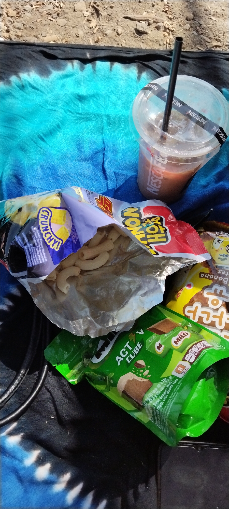 The image shows a close-up, top-down view of snacks and a drink placed on a blue and black tie-dye fabric. In the foreground, a bag of crunchy snack sticks is open, with several sticks visible inside. To the right of the snack bag, a clear plastic cup with a black straw contains a brown beverage, possibly iced coffee, with a Nescafé branded paper band around the lid. Partially visible in the lower right corner is a green package of Milo Active Cube, and behind it, a yellow package with Korean text. The fabric appears to be laid out on a sandy or rocky surface, suggesting an outdoor setting. The scene is likely in Senggigi, Indonesia, given the presence of local snack packaging. There are no people visible in the image.