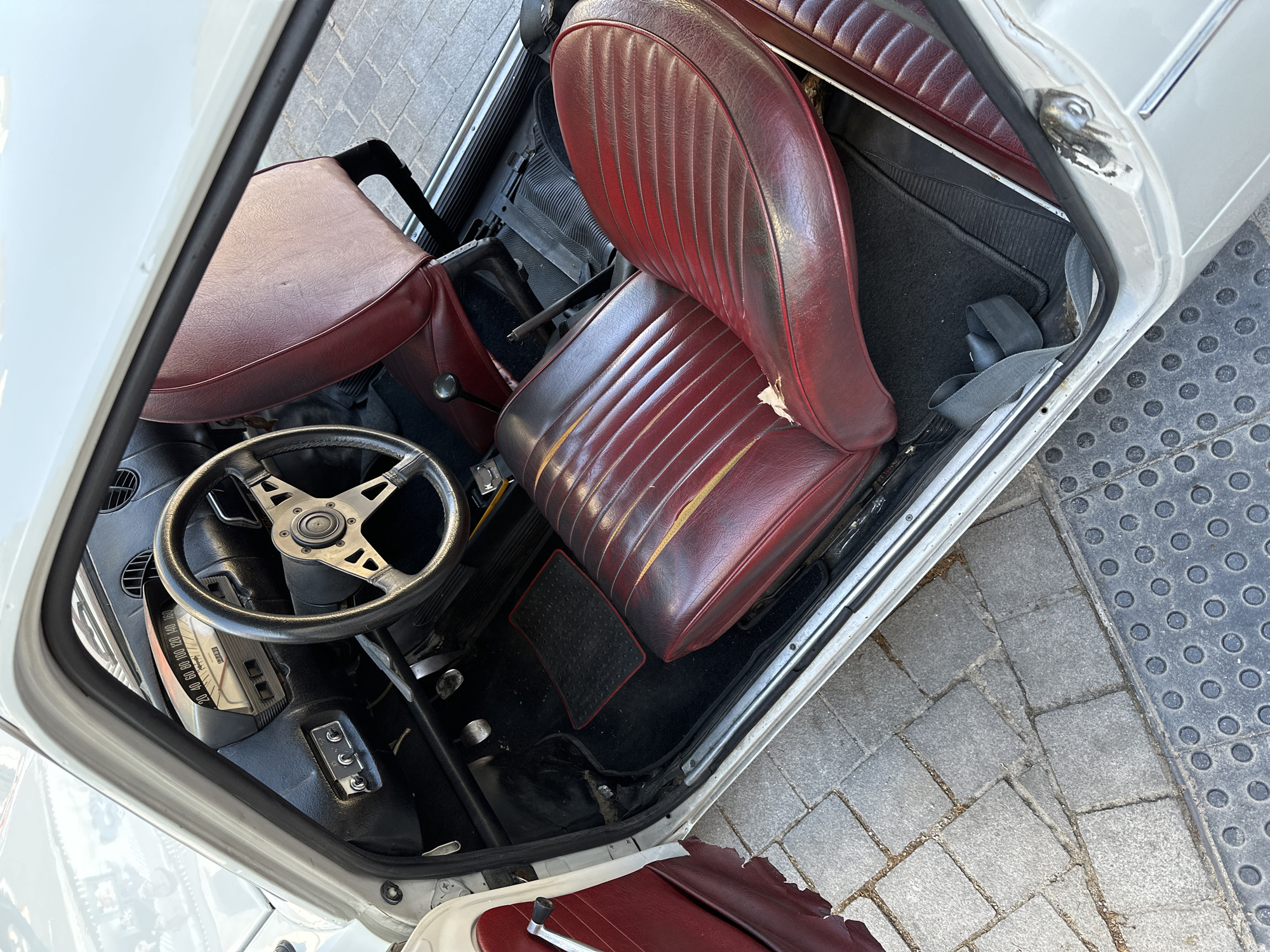 The image depicts the interior of a vintage car, specifically the driver's side. The seats are upholstered in a worn, burgundy-colored material with a ribbed pattern. The steering wheel has a four-spoke design and is connected to a dashboard with gauges and switches. The floor is black with a small, red-bordered floor mat visible. The car's door is open, revealing a glimpse of the exterior, which is a light color. The background shows a paved surface, indicating the car is likely parked outdoors. There is no evidence to confirm this location is in Madrid, Spain. No people are visible in the image. The focus is on the car's interior, highlighting its vintage condition.