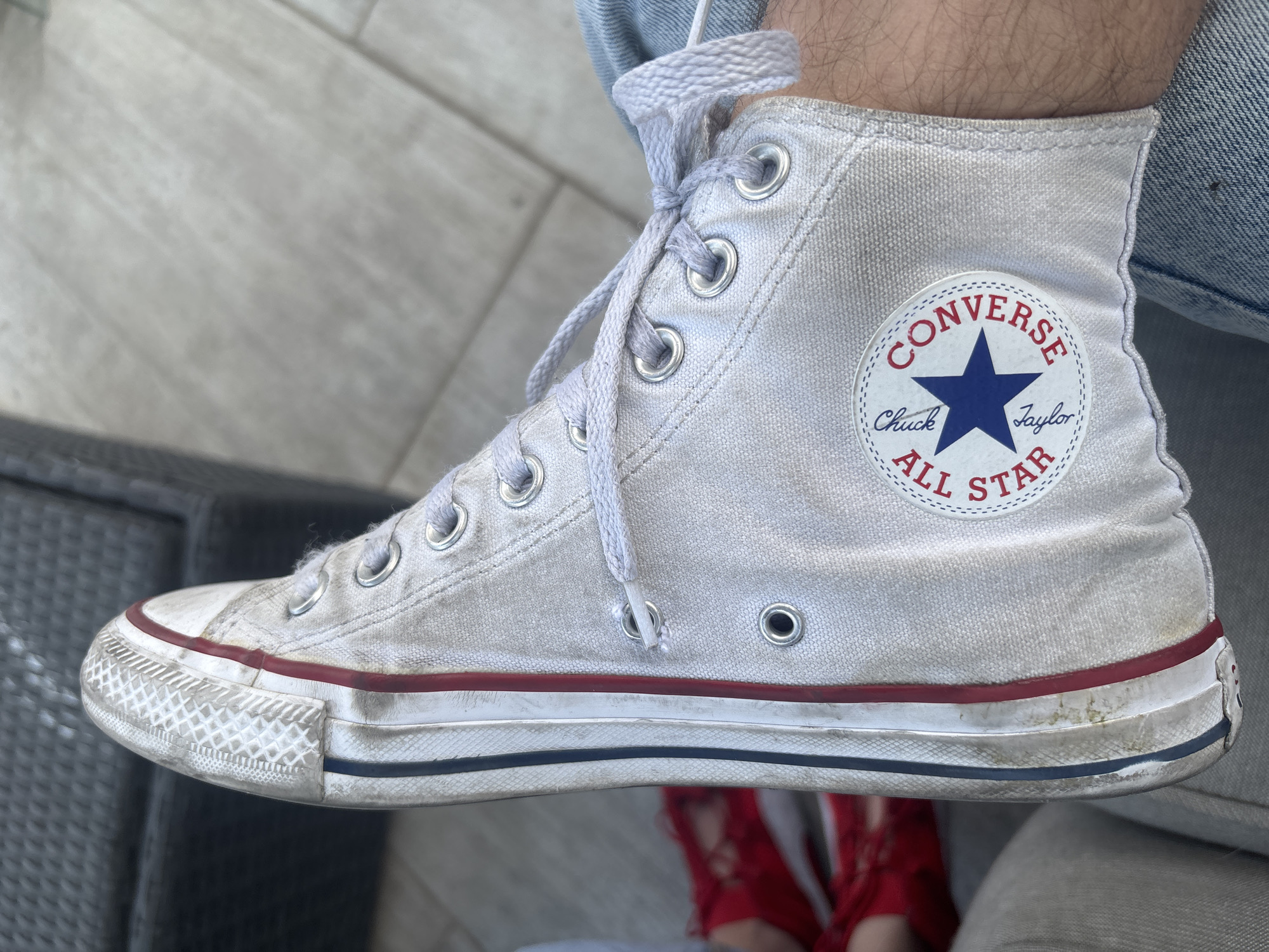 A close-up shot depicts a worn, white Converse Chuck Taylor All Star high-top sneaker. The shoe is laced up, but the laces are untied and hanging loosely. The iconic circular Converse logo with a blue star is visible on the side of the shoe. The sole of the sneaker is dirty and features the signature red and blue stripes. The person wearing the shoe is also wearing blue jeans. In the background, red shoes are visible. The photo appears to have been taken outdoors. I cannot determine a location in the city of Huixquilucan de Degollado, Mexico from the image.