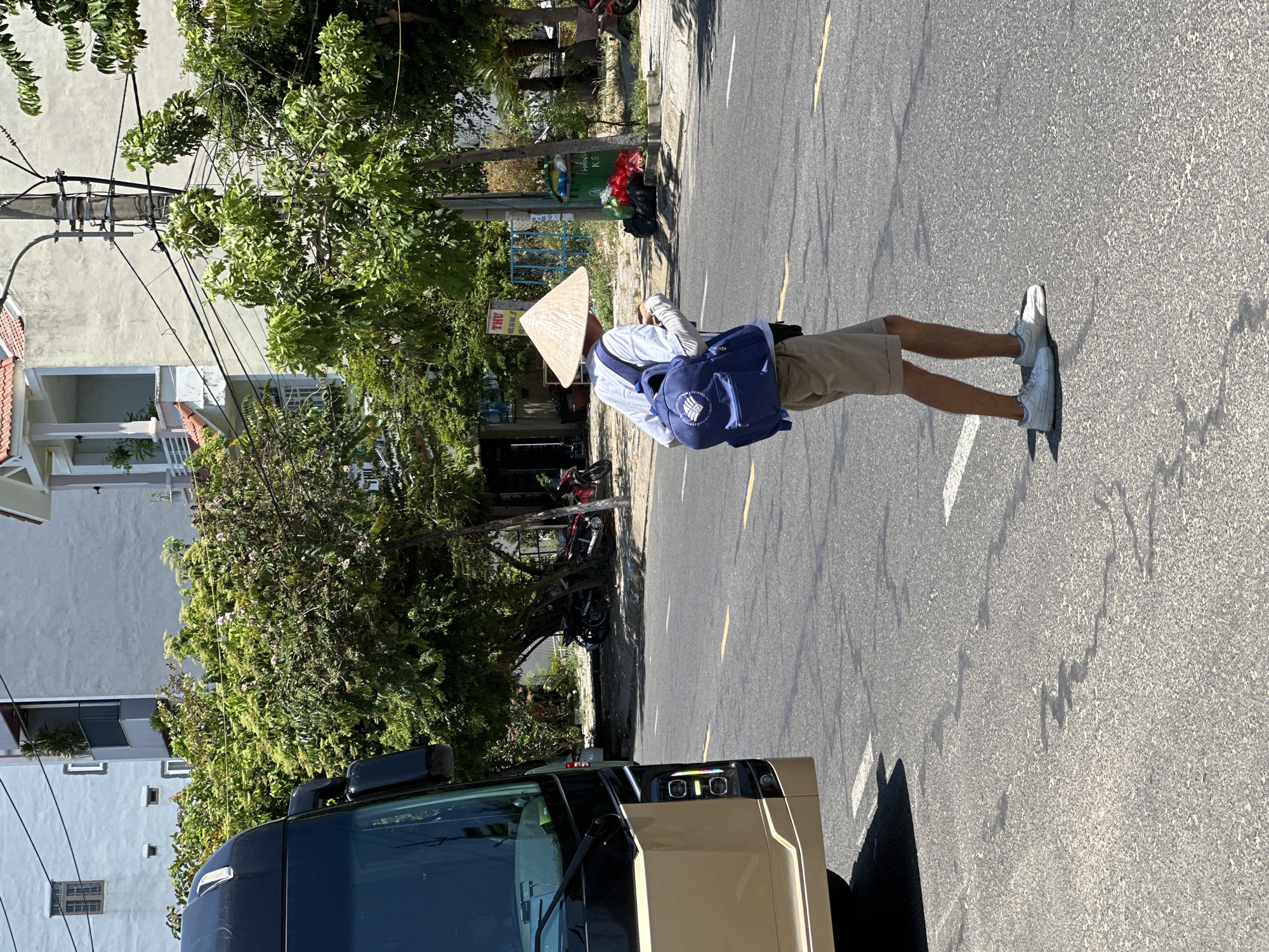 In Đà Nẵng, Vietnam, a person wearing a conical hat, a white long-sleeved shirt, khaki shorts, and white sneakers walks across a paved road. They carry a blue backpack and appear to be looking at their phone. A golden bus is parked on the left side of the road. The background shows buildings, trees, and parked motorcycles. The sky is clear and bright, indicating a sunny day.