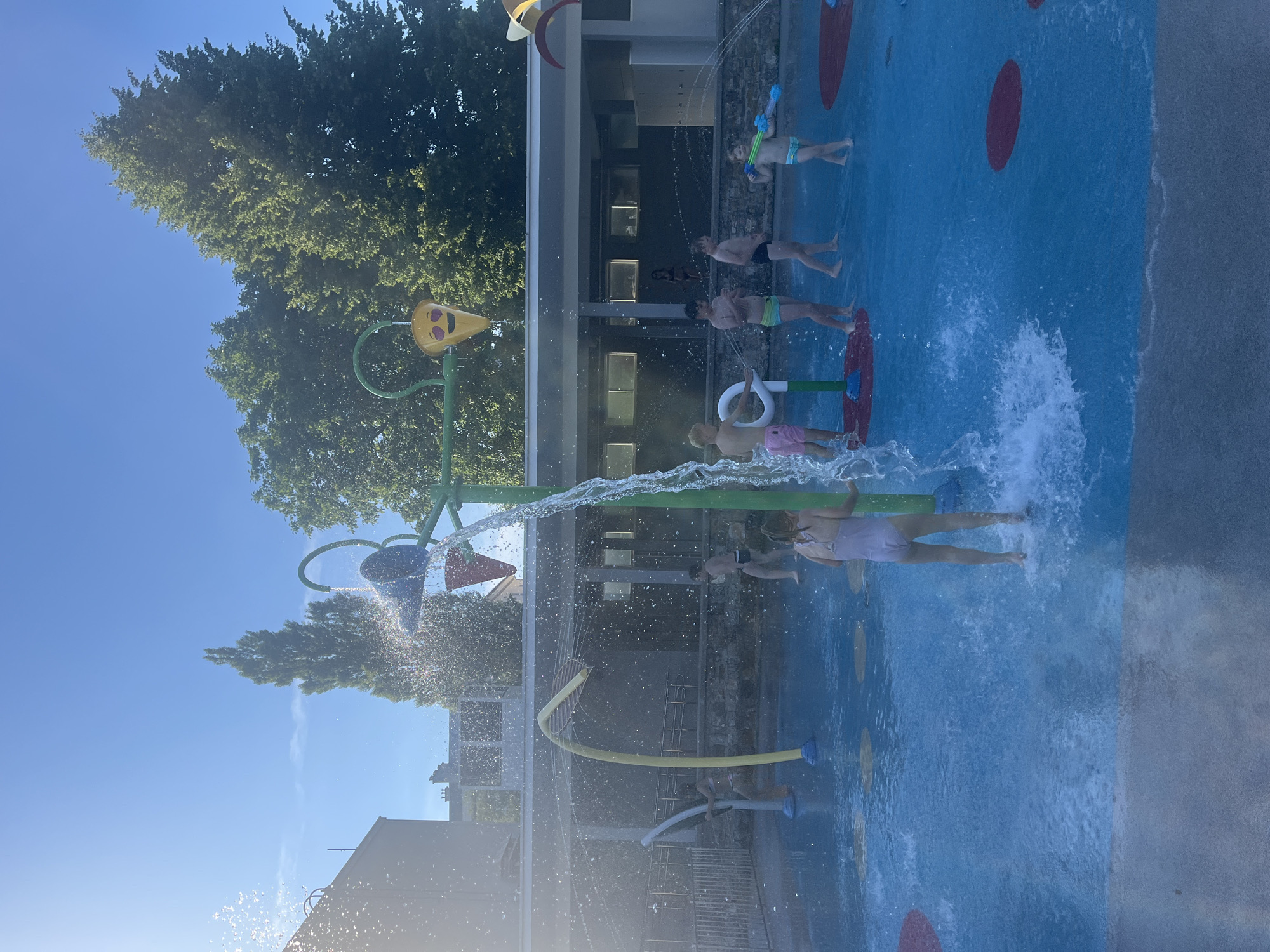 The image depicts a busy outdoor splash pad on a sunny day. Several children are actively playing in the water, with one child in the foreground wearing a pink swimsuit and splashing water. Other children are visible throughout the scene, some holding water toys and others interacting with the water features. The splash pad has a bright blue surface with red circular markers. Various water-spraying structures are present, including a large green structure with a bucket at the top that appears to be tipping water. In the background, there is a building with a colonnade and trees. The sky is clear and blue, indicating fair weather.