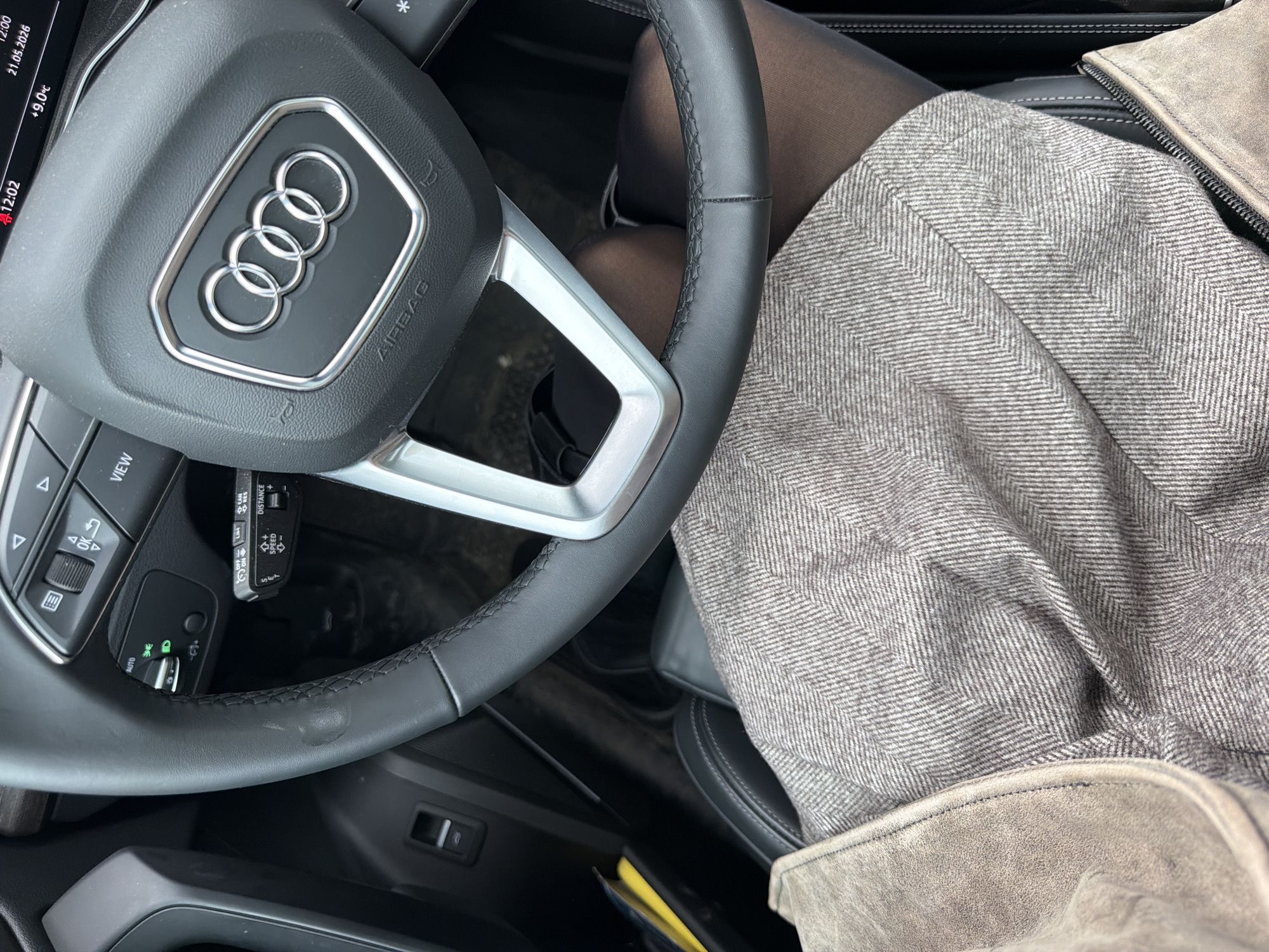 The image shows the interior of an Audi car, focusing on the steering wheel, part of the driver's seat, and a portion of the driver's attire. The steering wheel features the Audi logo and various controls. A leg clad in dark tights is visible, as is a garment made of grey herringbone fabric with a tan suede trim. The car's dashboard displays the time as 12:02, the temperature as +9.0°C, and the date as May 21, 2026. The photo provides no information to indicate its location in the city of Krasnoyarsk, Russia.
