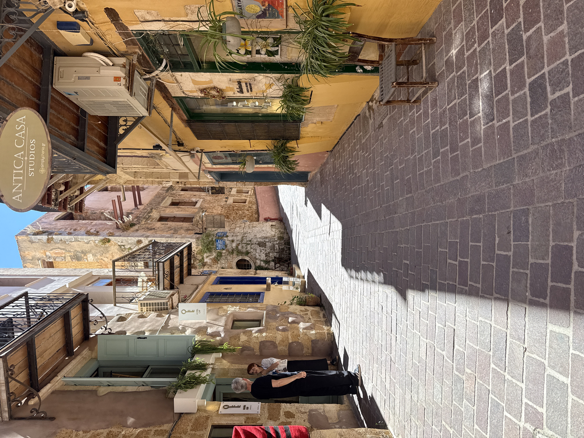 The image depicts a narrow, sun-drenched alleyway in Chania, Greece. The cobblestone street is paved with large, irregular stones. The buildings lining the alley are made of stone and plaster, with a mix of pastel and warm yellow hues.
On the left, a building features a light blue shuttered window and a balcony with ornate black metalwork. A white sign with a key symbol and the text "HOTEL OF" is visible. Two people, a man in a black t-shirt and black pants, and a woman in a white top and black skirt, stand near the entrance of this building, appearing to converse.
On the right, the building is painted a vibrant yellow. It has dark green-shuttered windows and several potted plants, some hanging and some in window boxes. A sign above reads "ANTICA CASA STUDIOS" and "simplychania.gr". An outdoor air conditioning unit is mounted on the exterior wall. Further down the alley, a rustic wooden chair with a woven seat sits near the yellow building.
The alleyway recedes into the distance, with shadows cast across the cobblestones, indicating a bright, sunny day. The overall atmosphere is charming and evokes a sense of historical European streetscape.