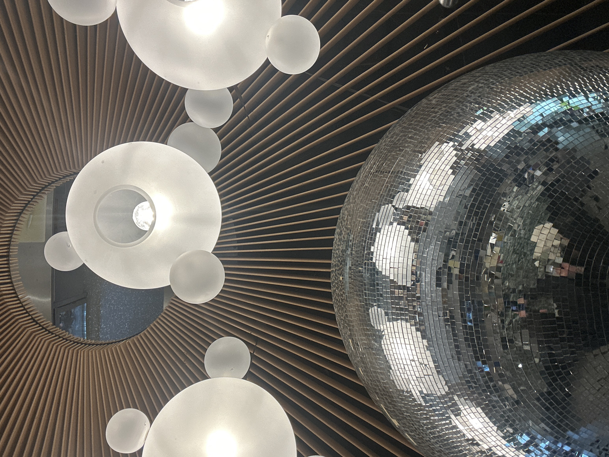 The image displays an interior ceiling view, likely within a commercial establishment in Varna, Bulgaria. The precise street address or specific venue within the city is not discernible from the visual information provided.
A large, spherical disco ball, composed of numerous small mirrored tiles, occupies the lower foreground, reflecting light and distorted elements of its immediate surroundings. Above this, the ceiling features a prominent radial design formed by parallel wooden slats, dark brown in color, converging towards a central circular opening. This opening reveals a darker, recessed area with a mottled or textured surface, possibly a vent or another structural element.
Scattered across the ceiling and interspersed within the wooden slat pattern are various illuminated light fixtures. These include multiple frosted white translucent globes of differing sizes and several larger, flat, circular frosted white translucent disc-shaped fixtures, each containing a visible incandescent bulb at its center. No people, actions, or interactions are present in the frame.