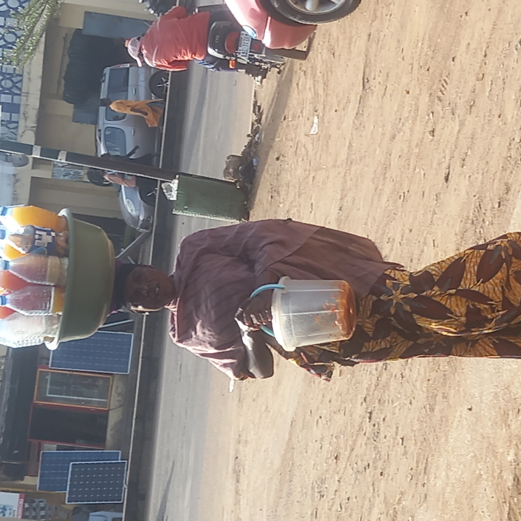 The image captures a street scene in Dadi Riba, Nigeria, featuring a woman engaged in street vending during daytime. The woman, who has dark skin, is prominently positioned, walking on an unpaved, sandy-colored roadside. She is dressed in a striped, dark-colored long-sleeved top and a patterned wrapper or skirt featuring brown, gold, and dark blue leaf-like designs.
On her head, she expertly balances a large, olive-green basin filled with numerous clear plastic bottles containing various colored beverages, including what appears to be juices and water. The label "H+" is discernible on one of these bottles. In her right hand, she carries a translucent plastic bucket with a light blue handle, holding a reddish-brown substance, likely a drink or food item. Her gaze is directed forward and slightly to her left, indicating focus on her path.
To the left of the woman, a paved road is visible, alongside which various other elements populate the background. A white van is present, and a person is seen on a red motorbike, whose license plate partially reads "LT-720C". Further back, several blue solar panels are visible on the ground or leaning against a structure, suggesting an area with commercial activity or makeshift setups. A building or wall with a distinct blue and white geometric pattern is also seen. The bright sunlight suggests clear weather conditions.