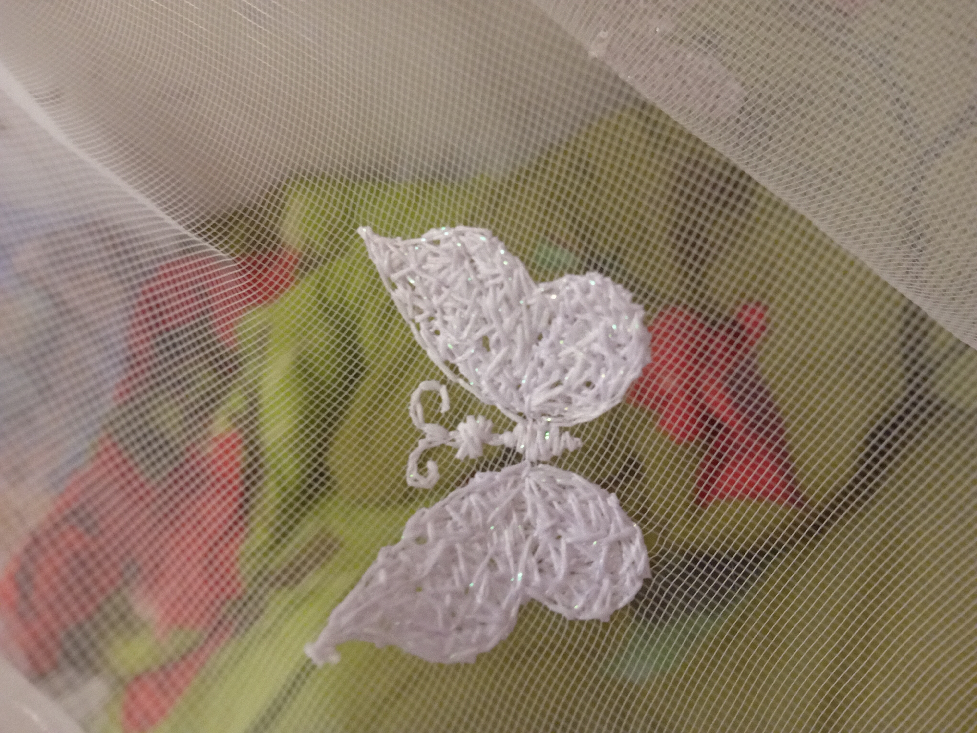 The image shows a white embroidered butterfly on a sheer white fabric. The fabric is draped, creating folds and a textured surface. In the background, there is a blurred image of red and green elements, possibly flowers or foliage. The image appears to be a close-up shot, with a shallow depth of field that blurs the background. The location in the city of Chuhuiv, Ukraine, cannot be determined from the visual information provided in the image.