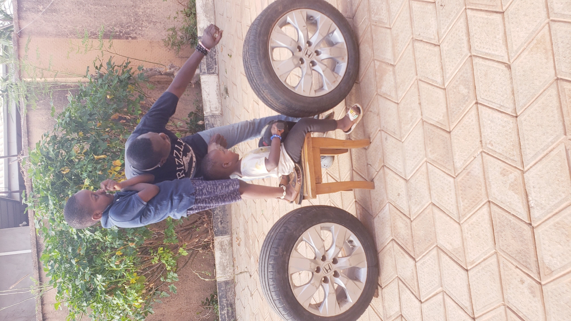 This outdoor image, taken in Sabon Tasha, Nigeria, features an adult male interacting playfully with two young children. The scene is set on a light-colored, diamond-patterned paved surface, with lush green foliage and a light brown wall forming the background.
On the left, an adult male, wearing a dark t-shirt with "RIZZLI" text and grey jeans, stands or leans slightly, with his right arm raised, fist clenched, in a gesture that suggests strength or celebration. He also wears beaded wristbands. In front of him, a young boy in a blue t-shirt and patterned shorts looks up at the adult, seemingly engaged in the interaction.
To the right, a younger child sits on a small wooden stool. This child wears a light-colored top, dark leggings, and sandals, and also has wristbands. They are looking towards the adult and the other child, holding a small object in their hands.
Two car wheels, both featuring silver five-spoke Honda rims, are prominently placed on the paved ground: one near the top right and the other towards the bottom right of the frame. The bright sunlight indicates it is daytime, and the overall atmosphere appears light-hearted and positive.