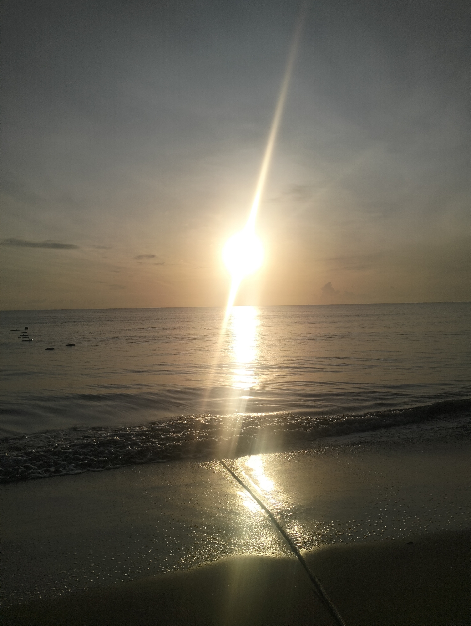 The image depicts a serene beach scene at sunrise in Calok, Indonesia. The sun is positioned high in the sky, emitting a bright, diffused light that casts a strong reflection across the calm ocean. Gentle waves lap at the shore, creating a foamy edge on the wet sand. A thick rope runs diagonally across the foreground from the bottom right corner, disappearing into the water. In the distance, several rectangular objects are visible, partially submerged in the sea. The sky is a soft gradient of grey and yellow, with hints of clouds. The overall atmosphere is peaceful and tranquil.