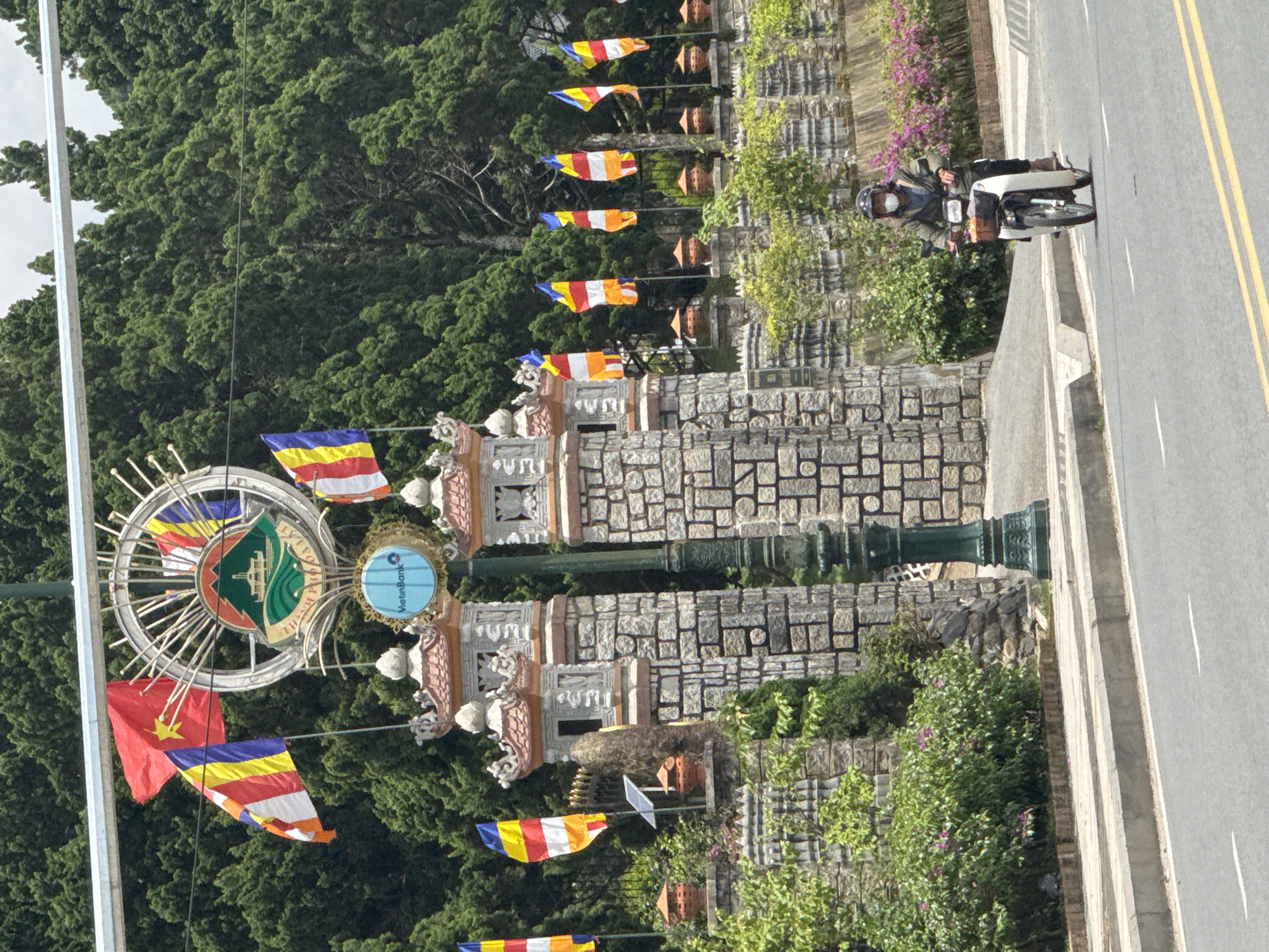 The image captures a view of the Thien An Pagoda in Da Lat, Vietnam. In the foreground, a person wearing a helmet and a face mask is riding a white scooter on a paved road. The scooter has a basket on the front containing a few items. In the background, there's a stone gateway with ornate towers and traditional Vietnamese architecture. Several colorful flags, including the Vietnamese national flag and Buddhist prayer flags, are displayed. Lush green trees and vegetation surround the pagoda, creating a vibrant and natural setting. The sky is partly cloudy.