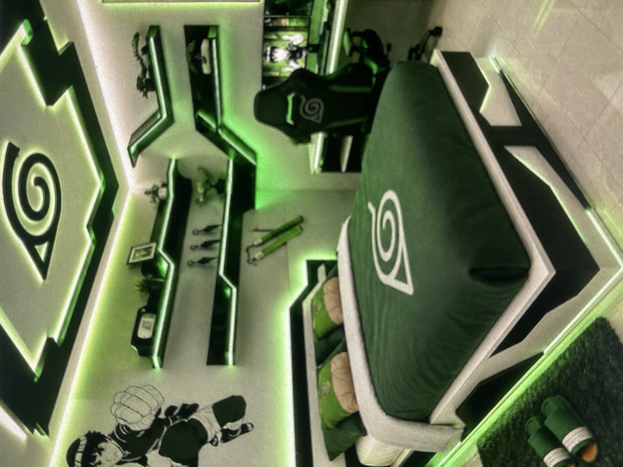 A room in Lhokseumawe, Indonesia, is decorated with Naruto-themed items. The walls feature stylized green and black designs with the Hidden Leaf Village symbol. The bed has green sheets adorned with the same symbol. Shelves display figurines and other Naruto-related objects. A gaming chair also incorporates the Hidden Leaf Village symbol. A rug sits on the floor near the bed.