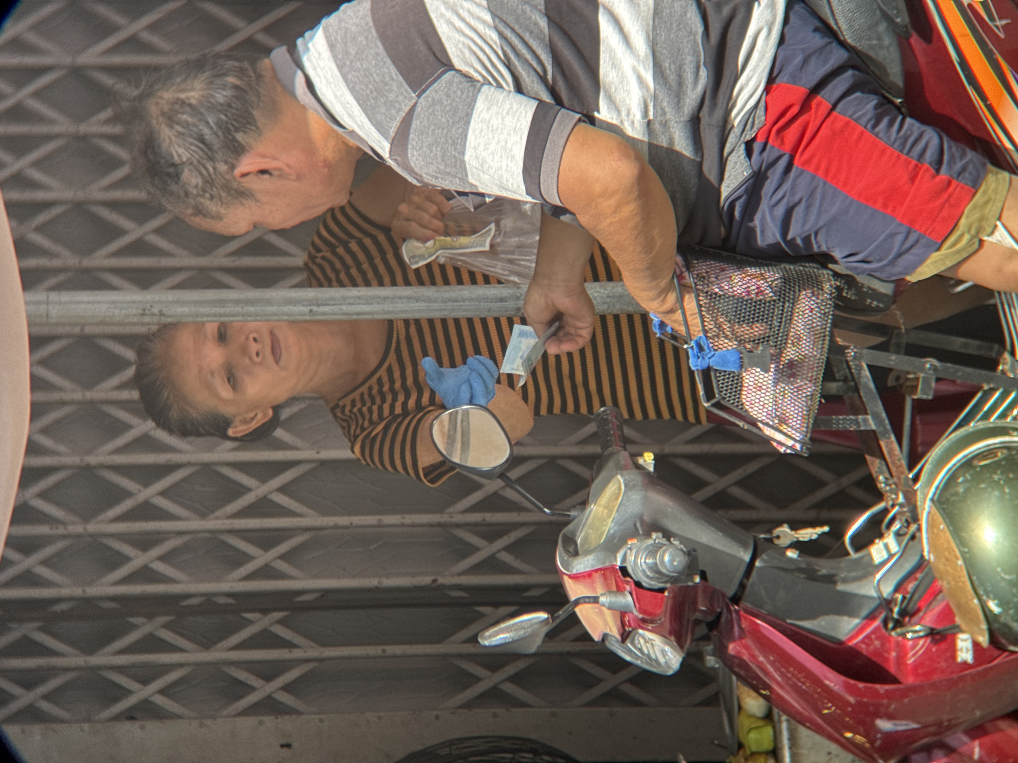 A transactional interaction occurs on a sunny day in Nha Trang, Vietnam, outside a commercial establishment with a metal roll-up gate. A man seated on a red and grey scooter, wearing a grey and white horizontally striped t-shirt and blue shorts with a red stripe, is receiving an item in a clear plastic bag from a woman. The woman, standing and wearing a black and orange horizontally striped t-shirt, holds the bag with her right hand. She is also handing a folded bill, possibly currency, to the man with her gloved left hand. The man is holding his right hand out, appearing to receive the bill. A black metal basket is attached to the front of the scooter, containing more items in plastic bags. The scooter has a red body, a silver front panel, and a green helmet is visible on the lower left. A metal pole stands between the man and the woman. The background consists of the closed metal gate.