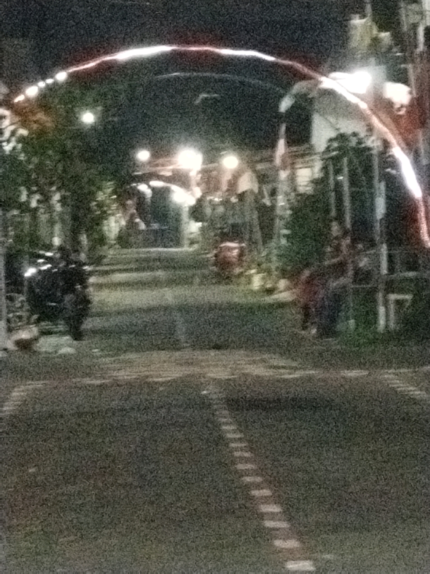 The image depicts a street scene at night, likely in a residential or commercial area of Watugede, Indonesia. A prominent archway adorned with lights spans across the road, casting a glow. Several motorcycles are parked along the sides of the street, one of which is in the foreground on the left. In the background, there appear to be buildings and trees, illuminated by streetlights and the decorative archway. The road is paved and marked with white lines, suggesting it is a pathway for vehicles. There are also hints of people in the background, possibly gathered or walking, but their details are indistinct due to the low light and image quality. The overall atmosphere is that of a lively evening in a town or city.