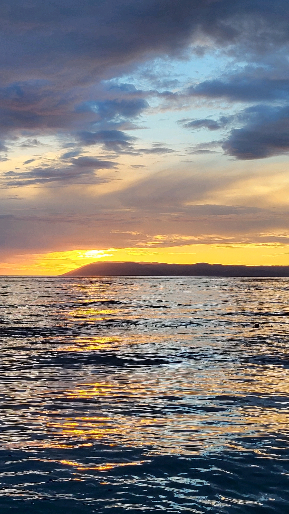The image depicts a sunset over the Adriatic Sea, likely near Općina Tučepi, Croatia. The sky is filled with a dramatic display of clouds, ranging in color from deep gray and blue to vibrant orange and yellow near the horizon where the sun is setting. The sun itself is partially obscured by a silhouetted mountain range in the distance. The sea in the foreground is calm, with gentle ripples reflecting the colors of the sunset. A series of small buoys or markers are visible in a line across the water. The overall impression is one of natural beauty and tranquility.